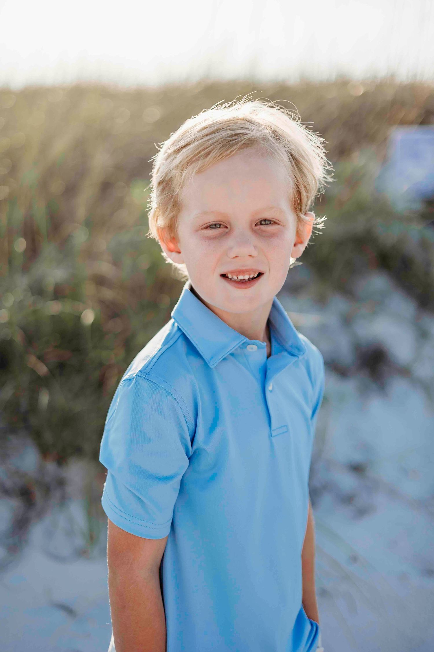 Young child in light blue polo shirt smiling on beach during golden hour.