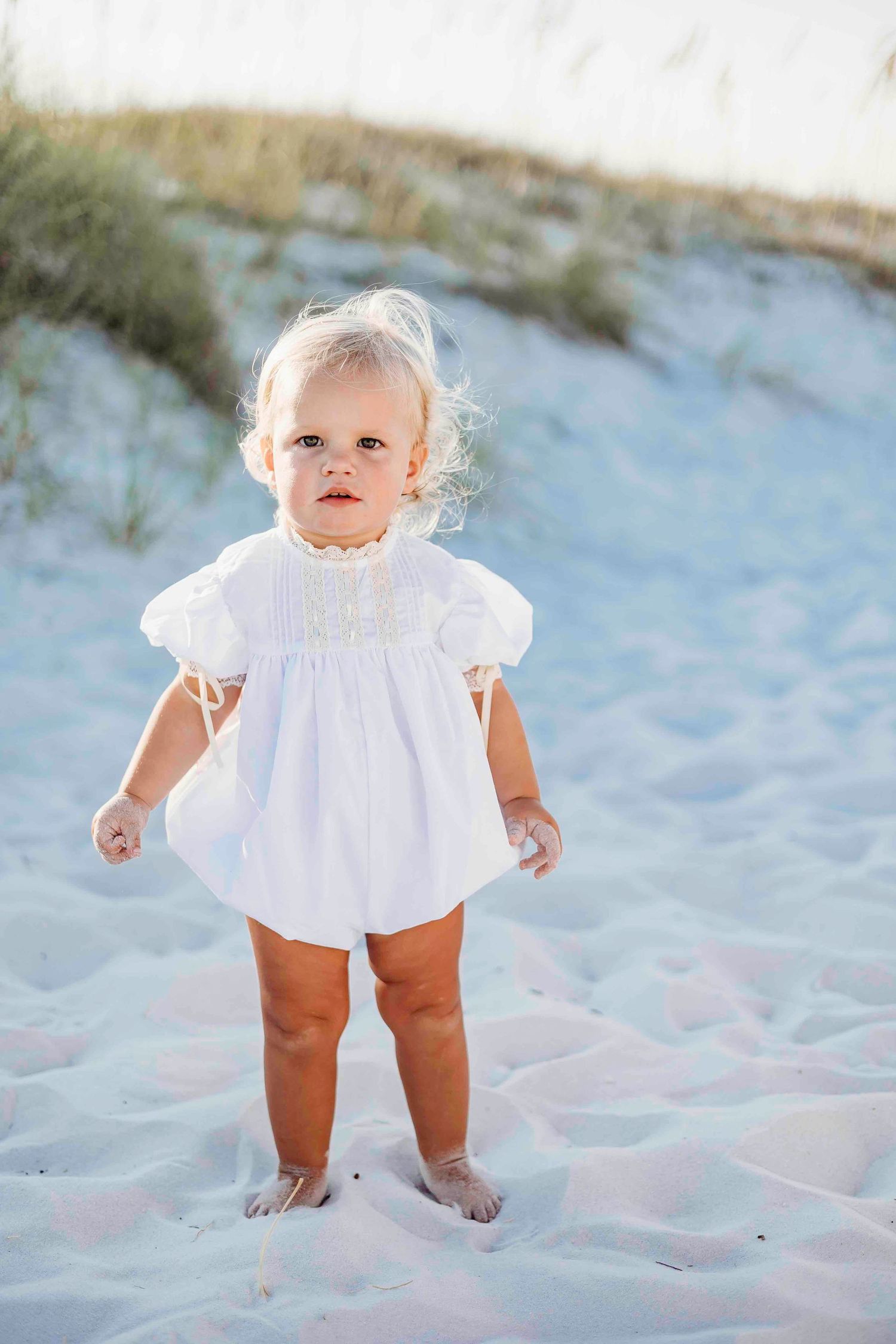 A toddler in white dress stands on white sand at the beach.