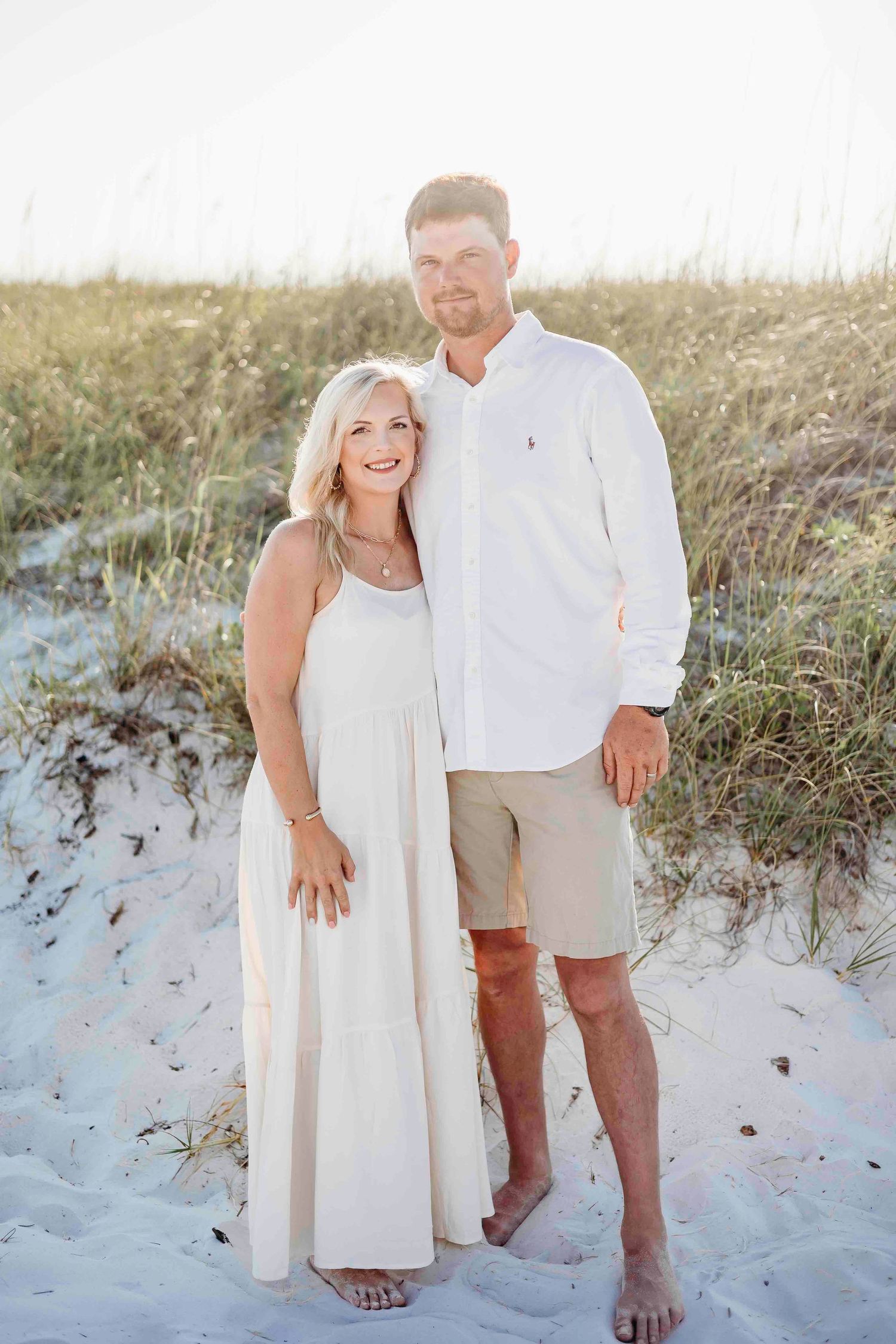 Couple in white beach attire posing together on sandy dunes at sunset.