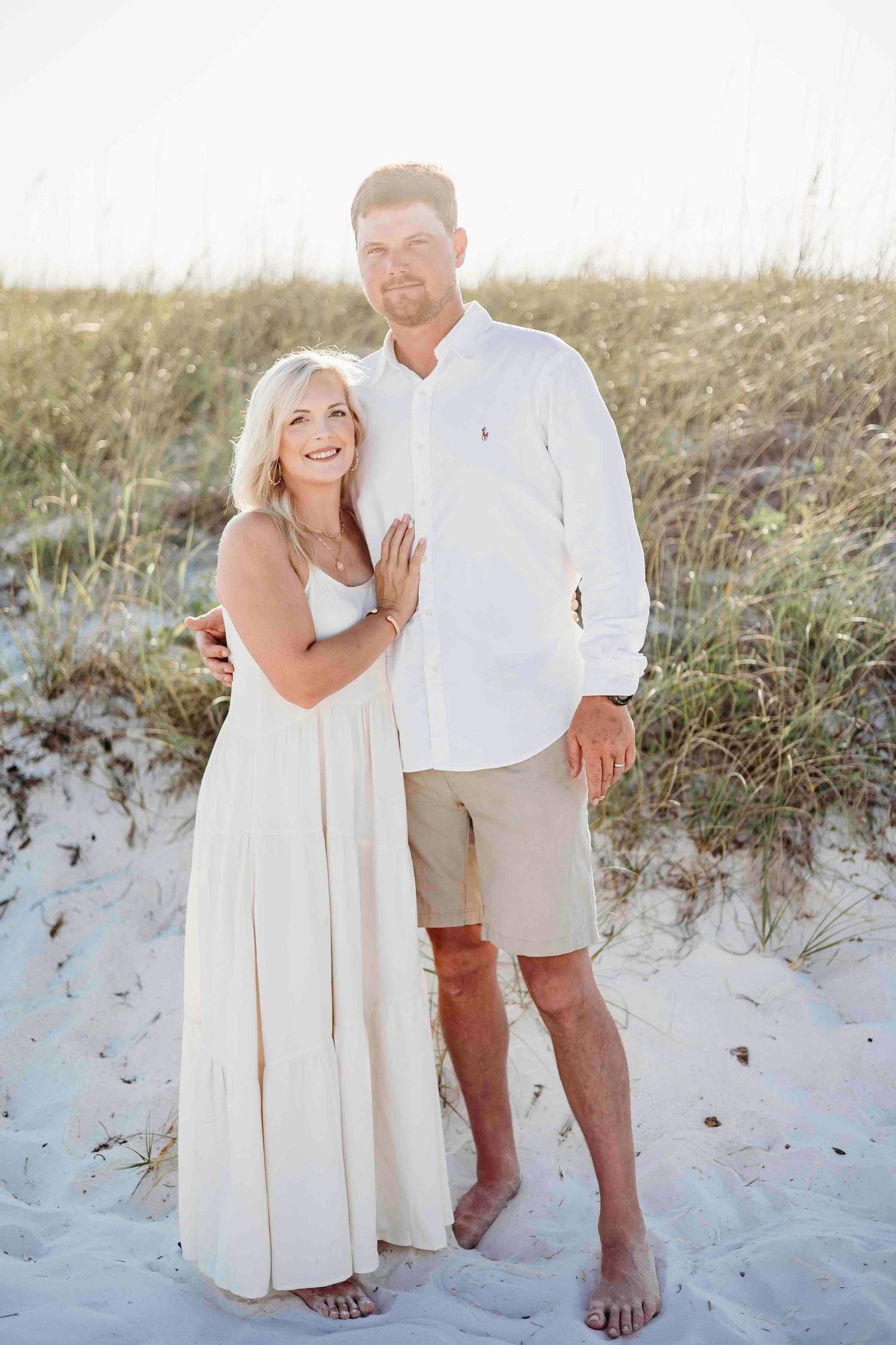 Happy couple in white and khaki beach attire standing barefoot on sand dunes.
