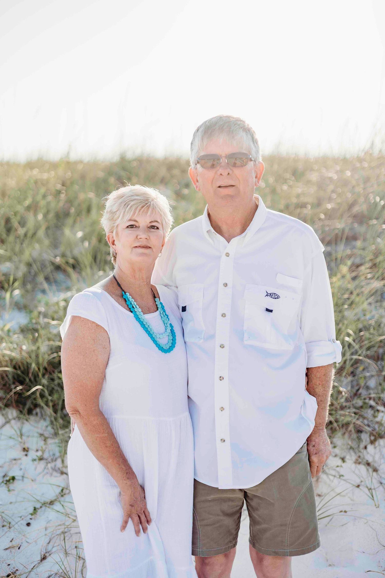 Mature couple in white attire and sunglasses posing on beach dunes.