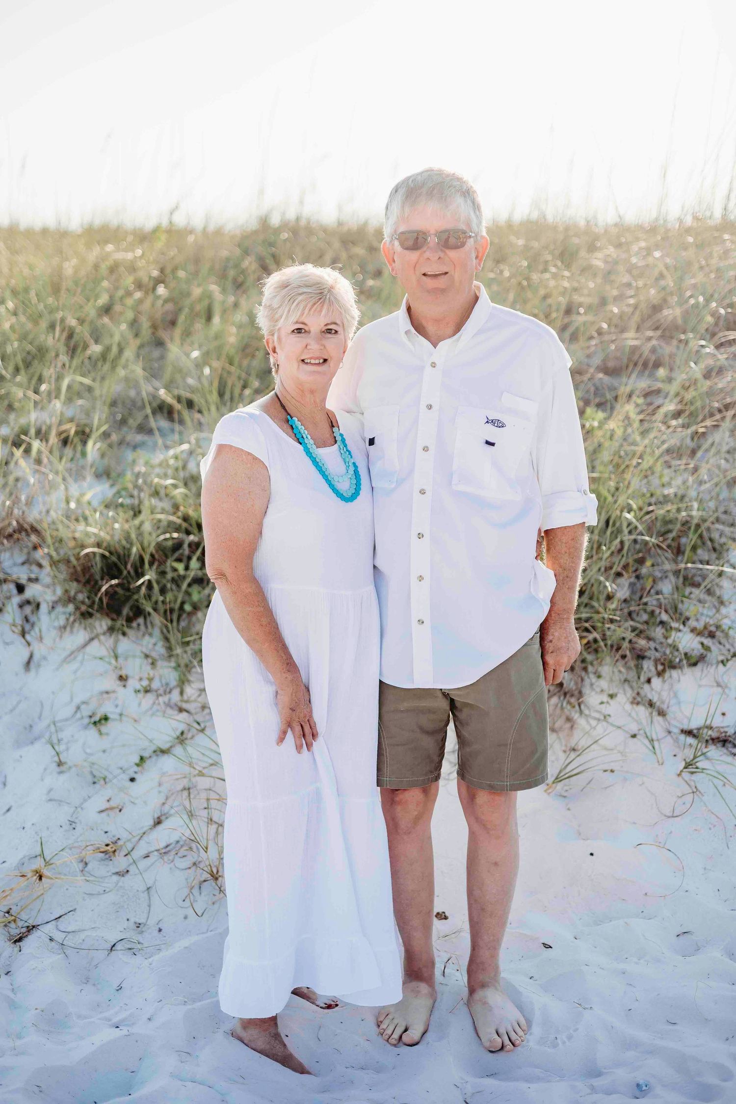 Senior couple in coordinated white outfits standing together on sandy beach.