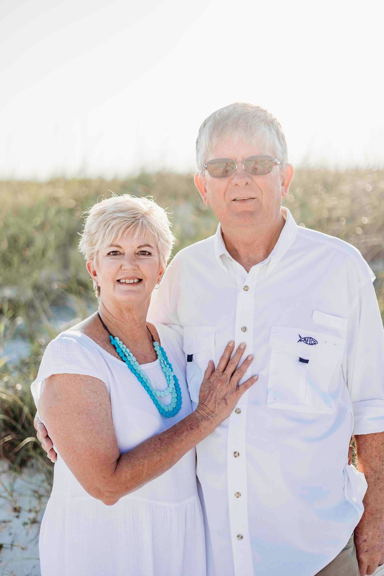 Loving senior couple in white clothing posing on beach with turquoise necklace accent.