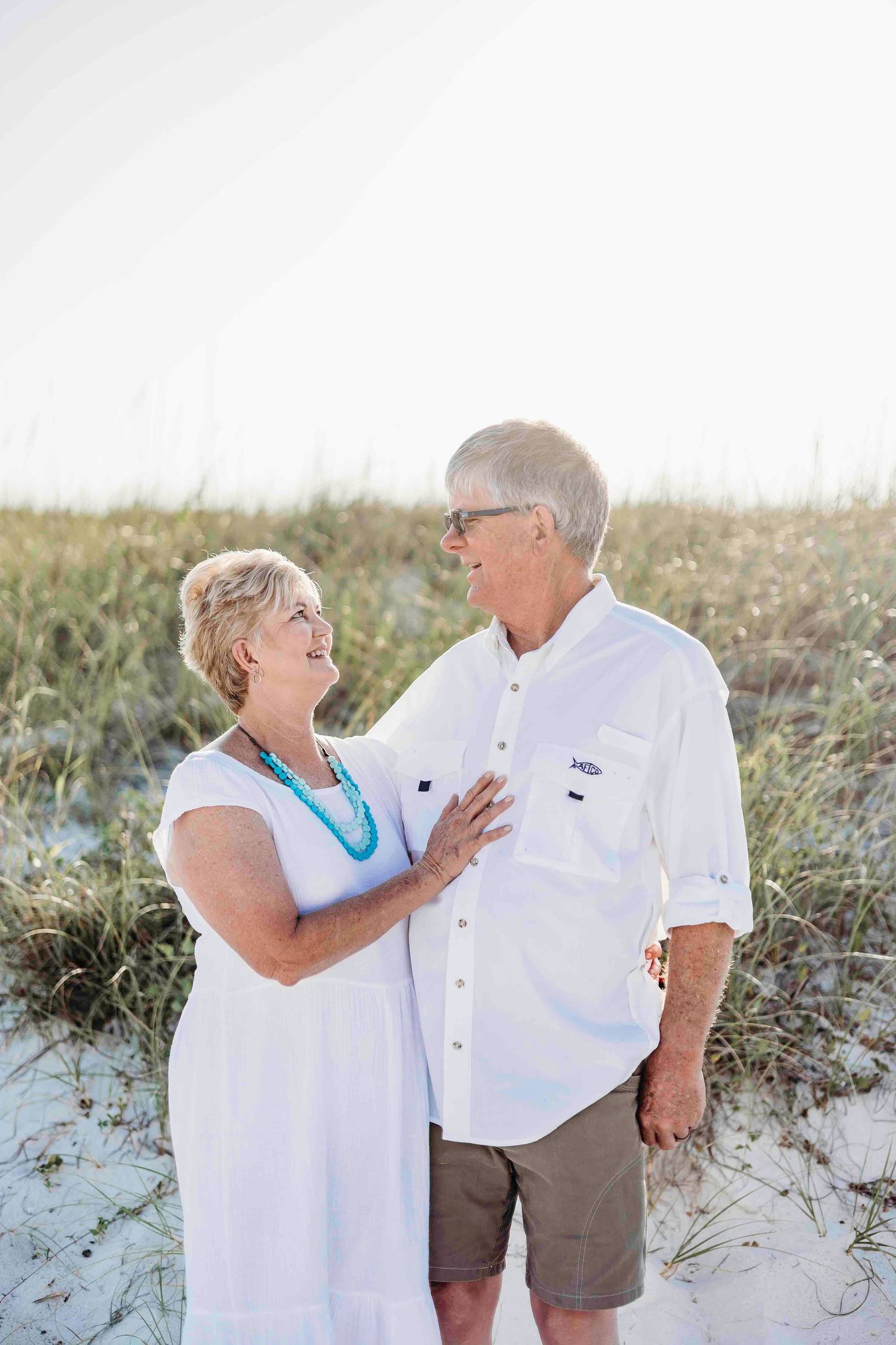 A couple wearing white outfits shares tender moments together on a sandy beach during sunset.