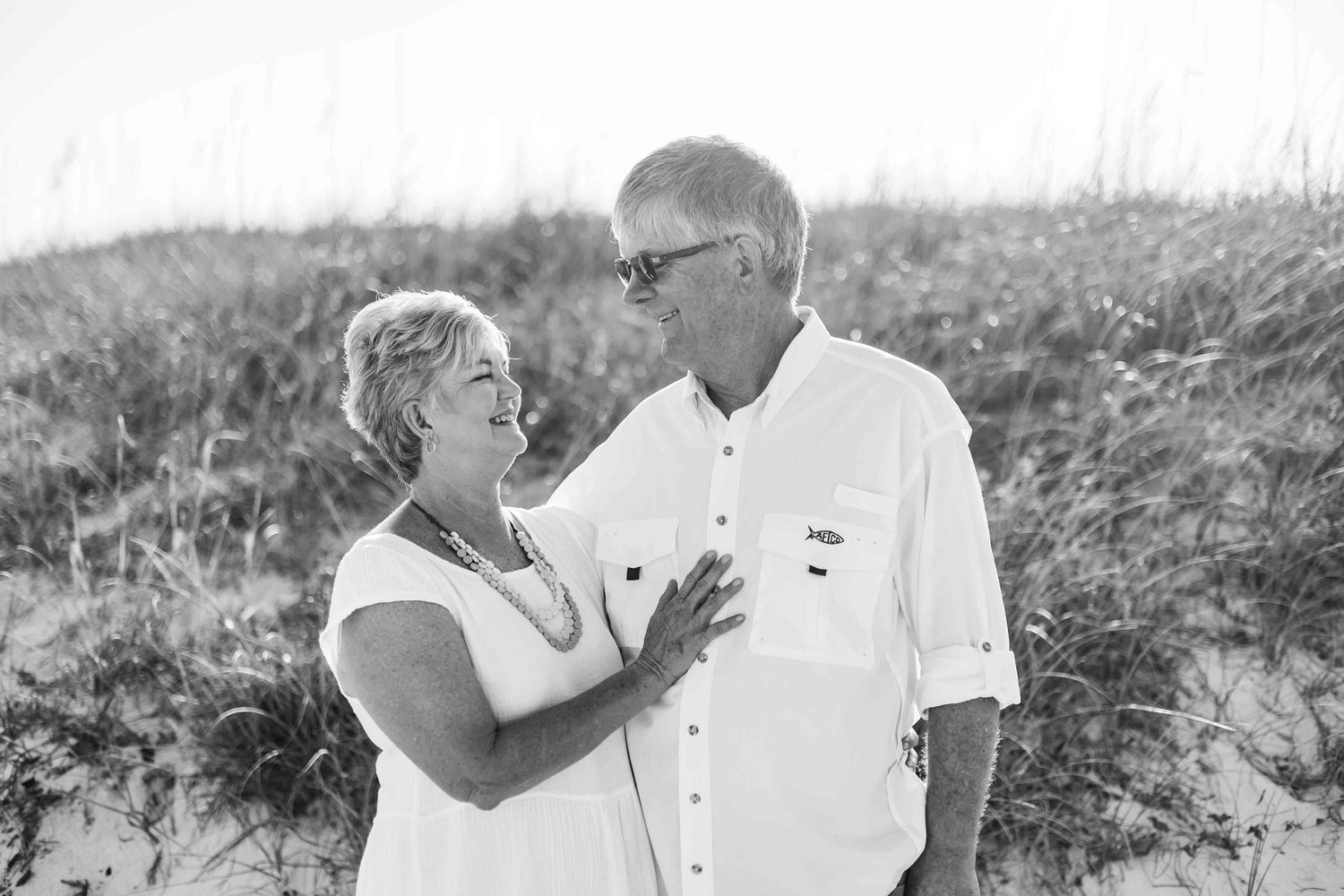 Romantic couple in white clothing embraces on beach dunes during a photo session.