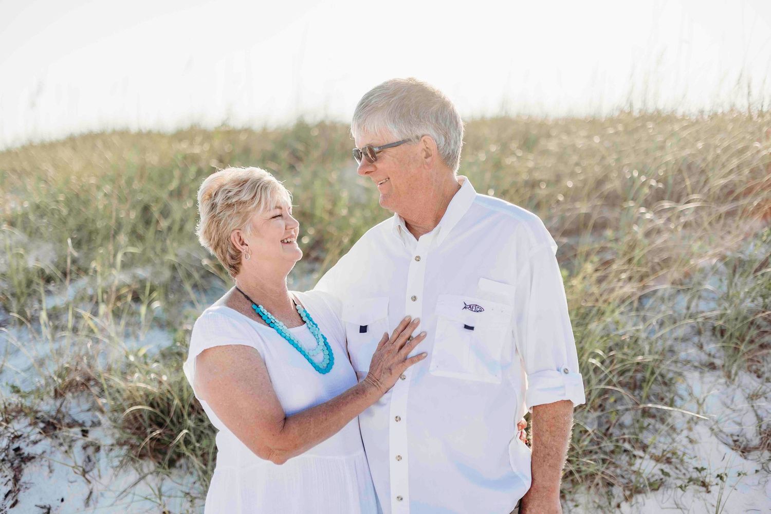 Senior couple in coordinating white outfits laughs together on coastal dunes.