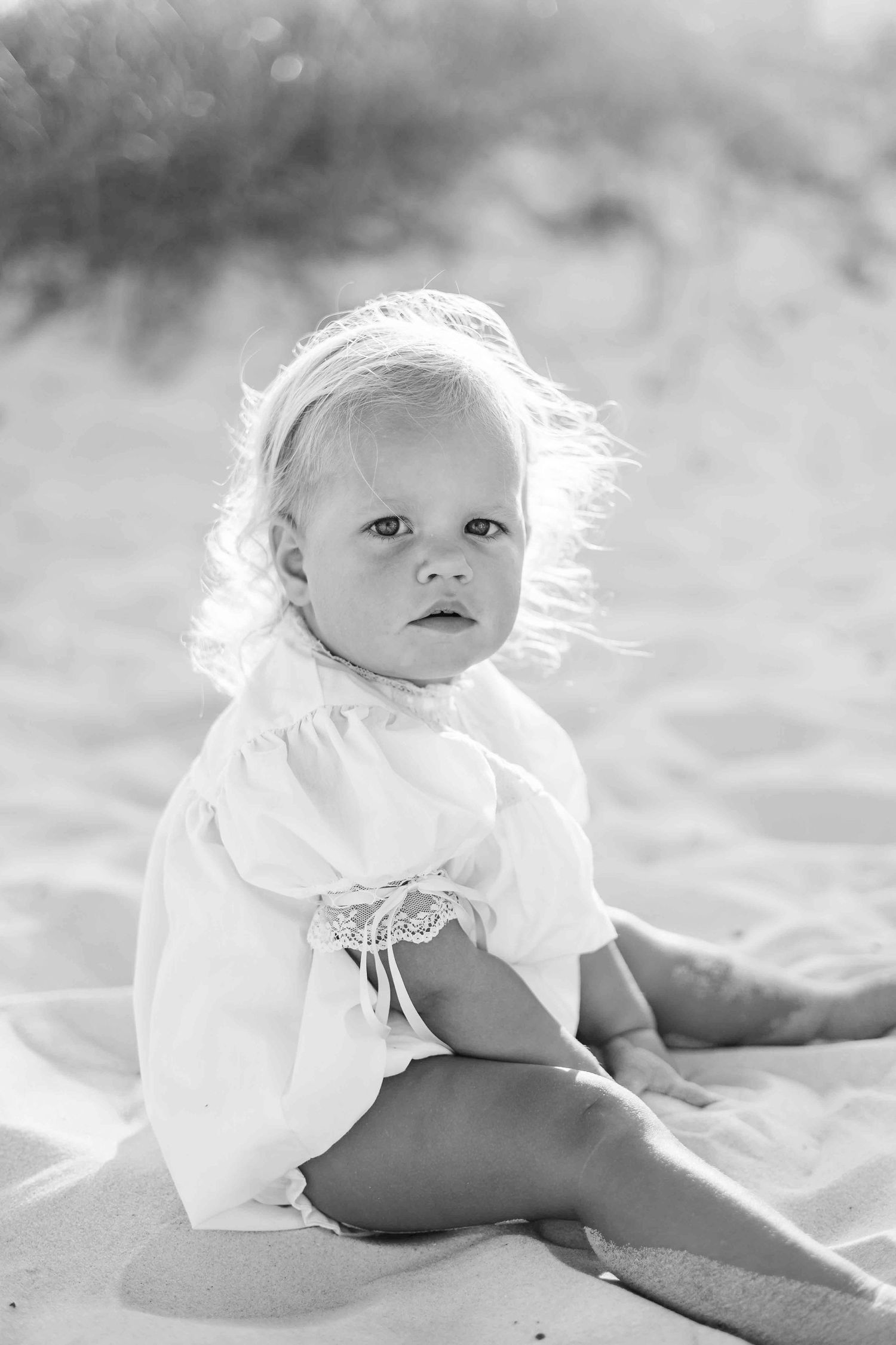 Baby in white dress poses on beach sand during sunset photo session.