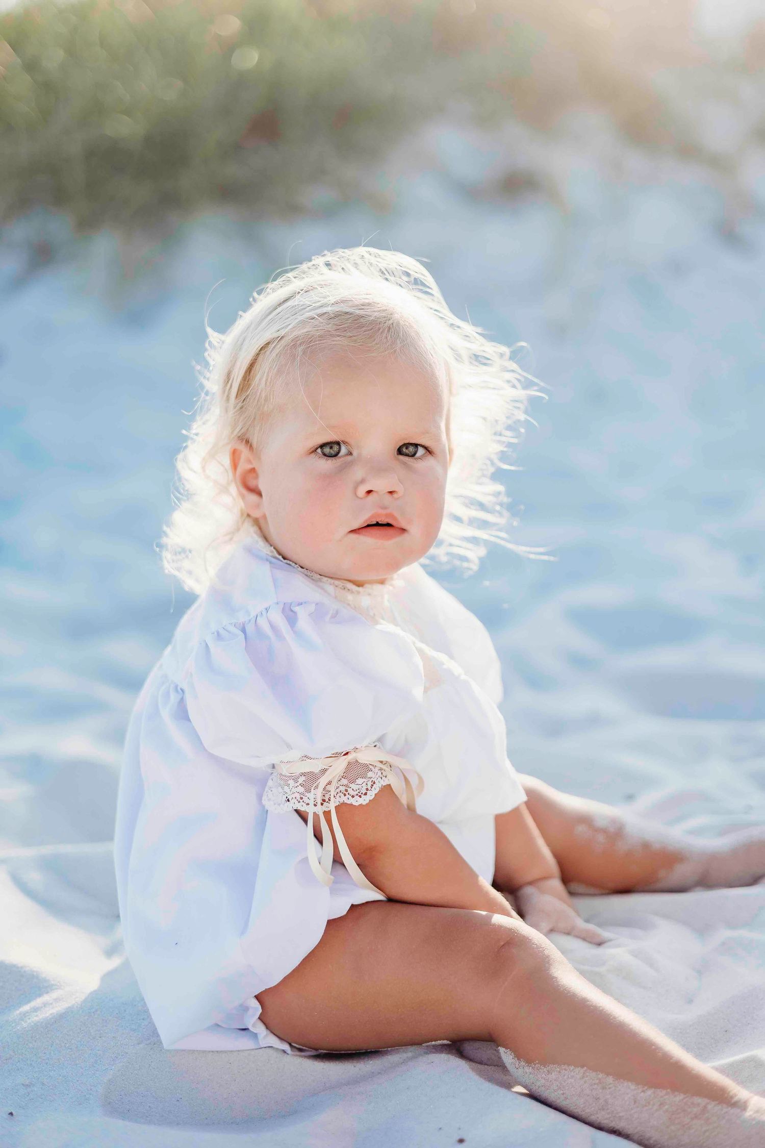Small child in white dress sits on sandy beach with windswept hair.