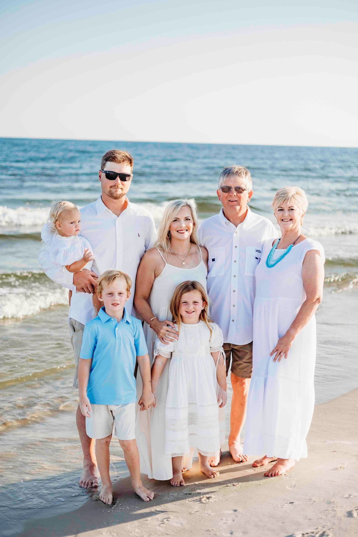 Multi-generational family wearing coordinated white and blue outfits poses together on sunny beach at sunset.