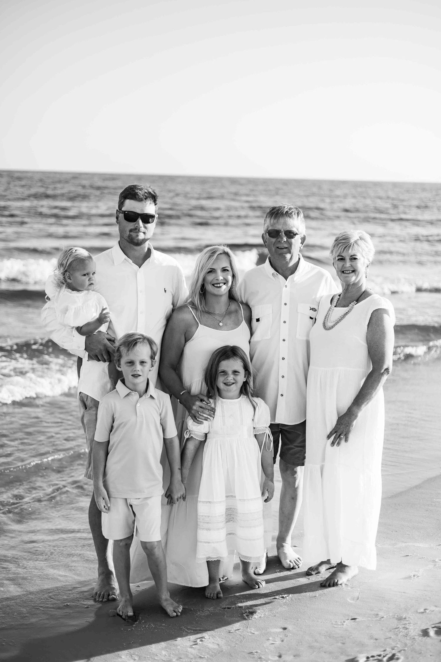 Black and white beach portrait of family group standing together by ocean waves.