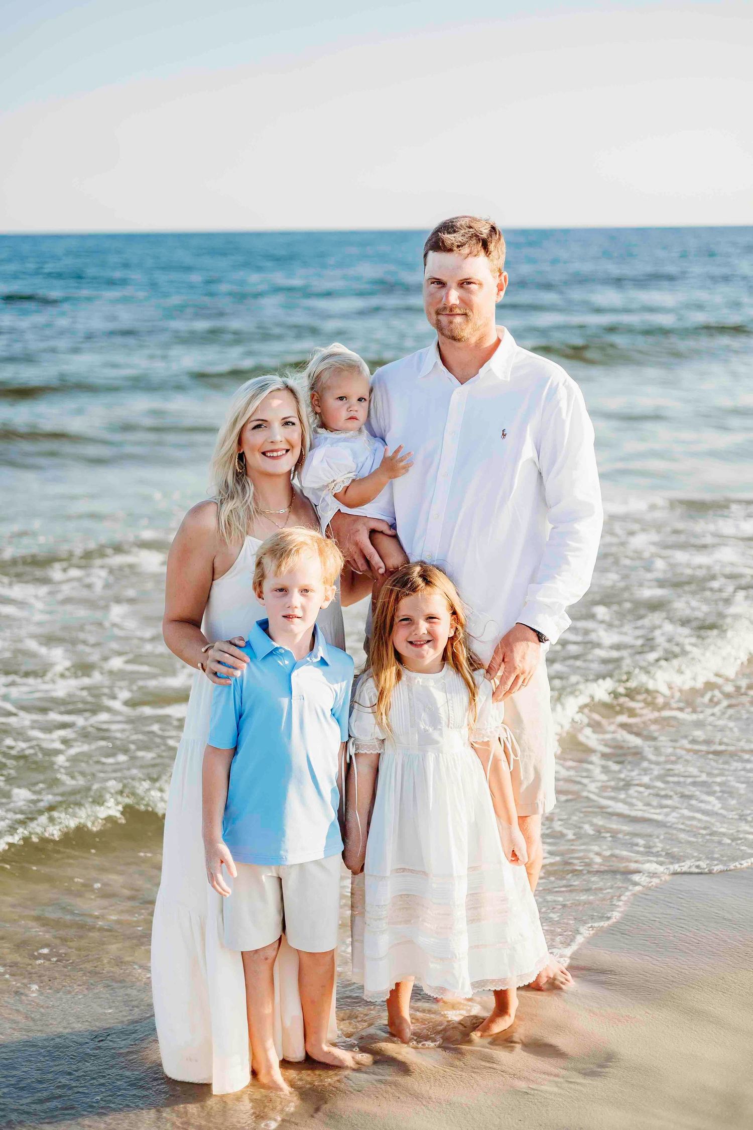 Extended family in casual beach attire smiles while posing together along shoreline at golden hour.