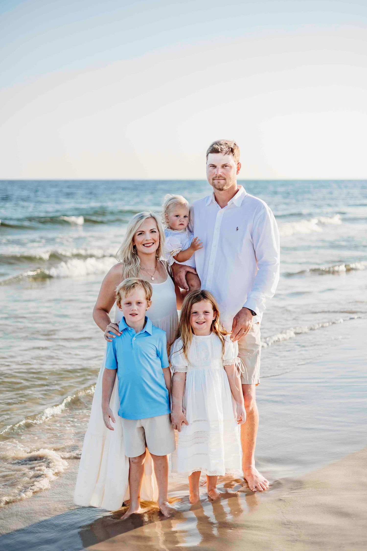 Family members in coordinated white and blue outfits gather for casual portrait on sandy beach.