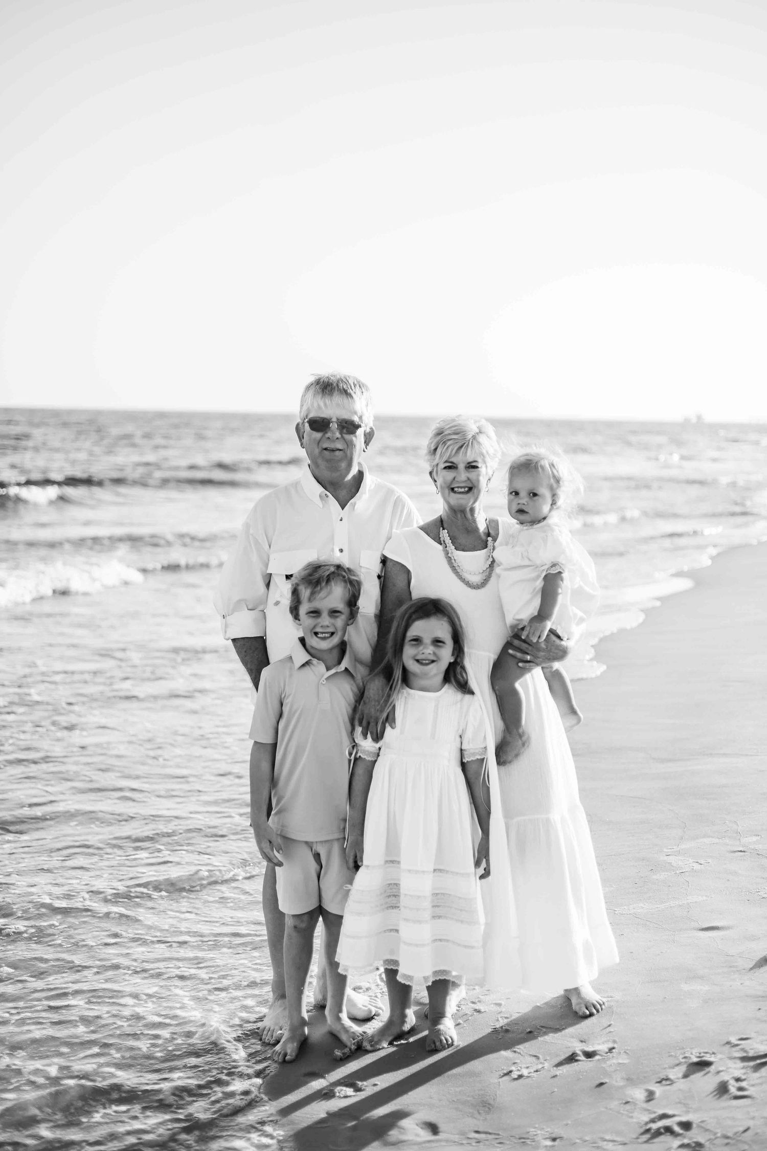 Black and white beach portrait showing family members gathered on sandy shoreline.