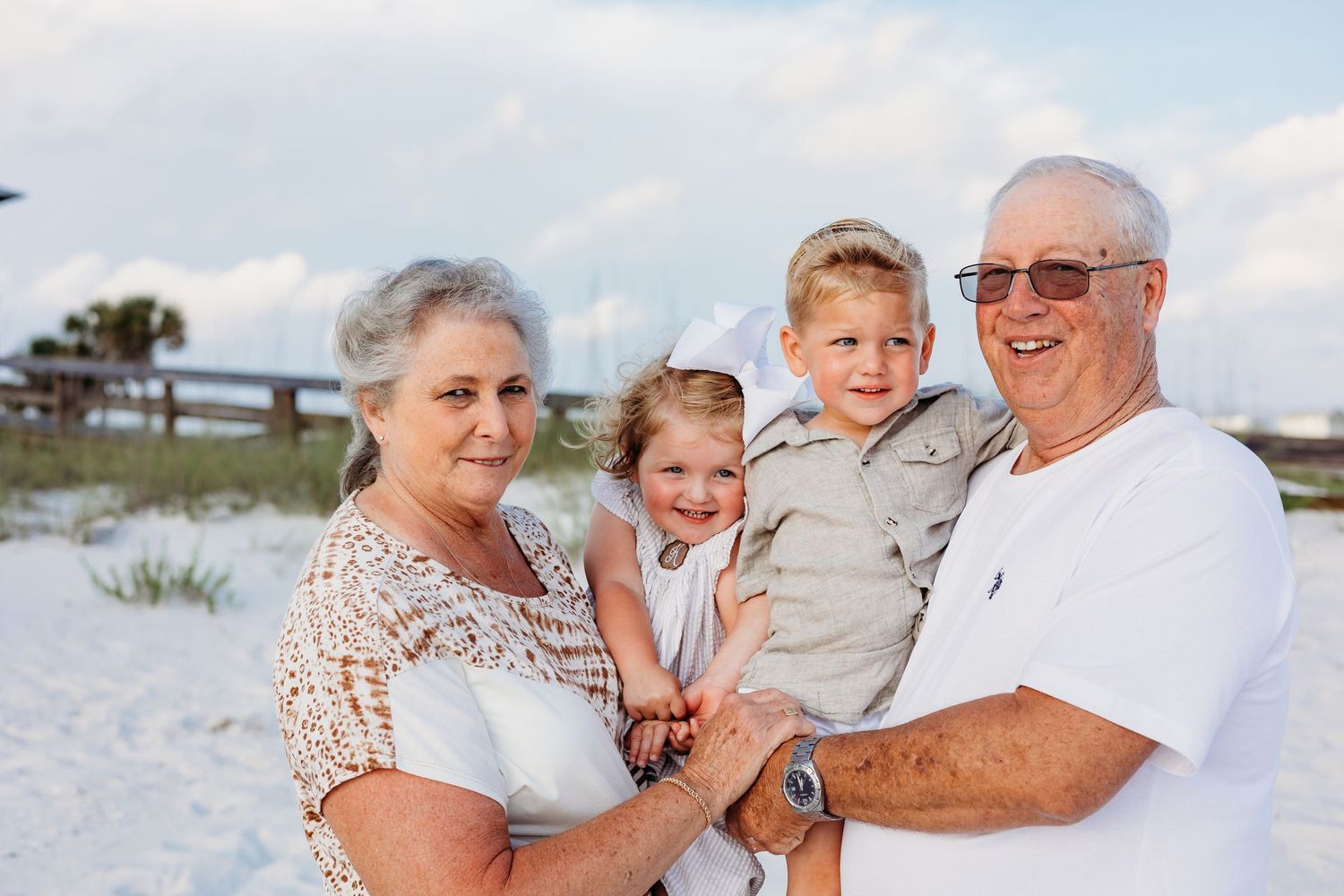 Happy family group poses together on white sandy beach with coastal vegetation in background.