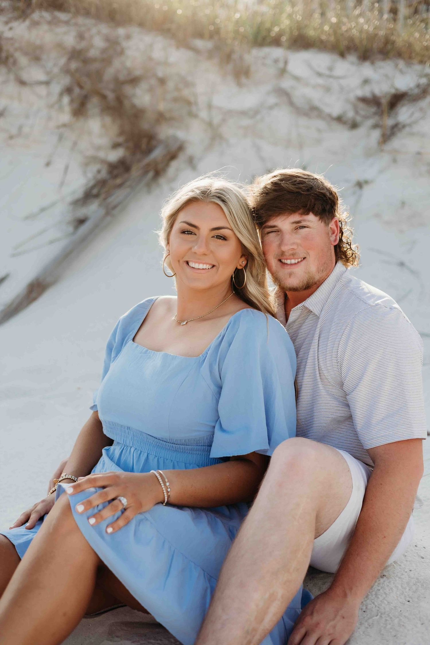 Couple poses intimately on beach sand wearing light blue dress and casual attire.