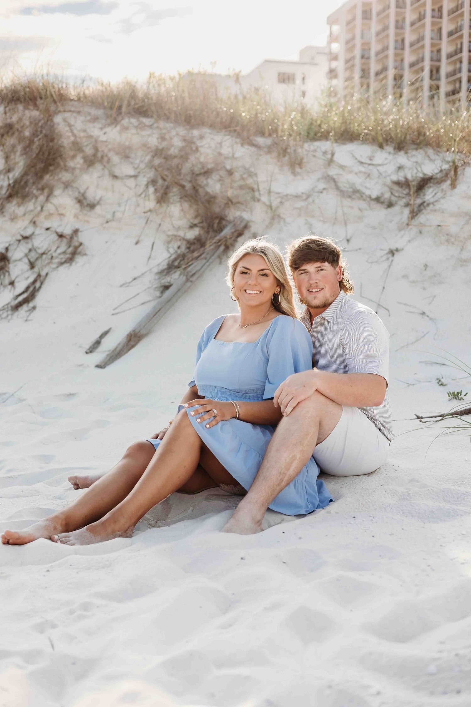 Couple enjoys romantic moment on sandy beach with coastal buildings in background.