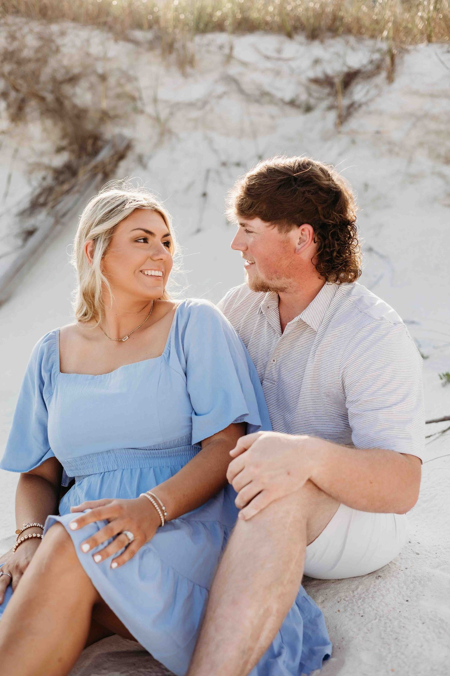 Couple sitting together on white sand beach sharing intimate moment in casual summer attire.