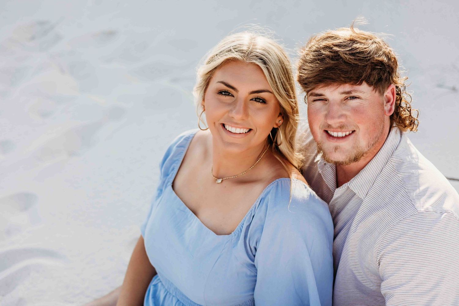 Romantic beach photoshoot with couple in light blue dress and white shirt against sandy backdrop.