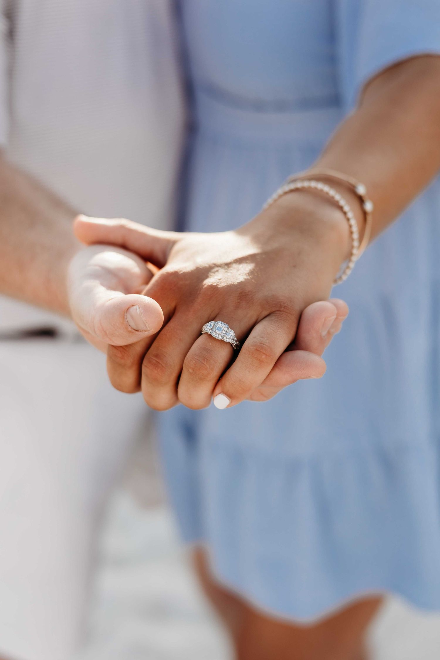 Sunlit close-up of hands with engagement ring against light blue fabric.