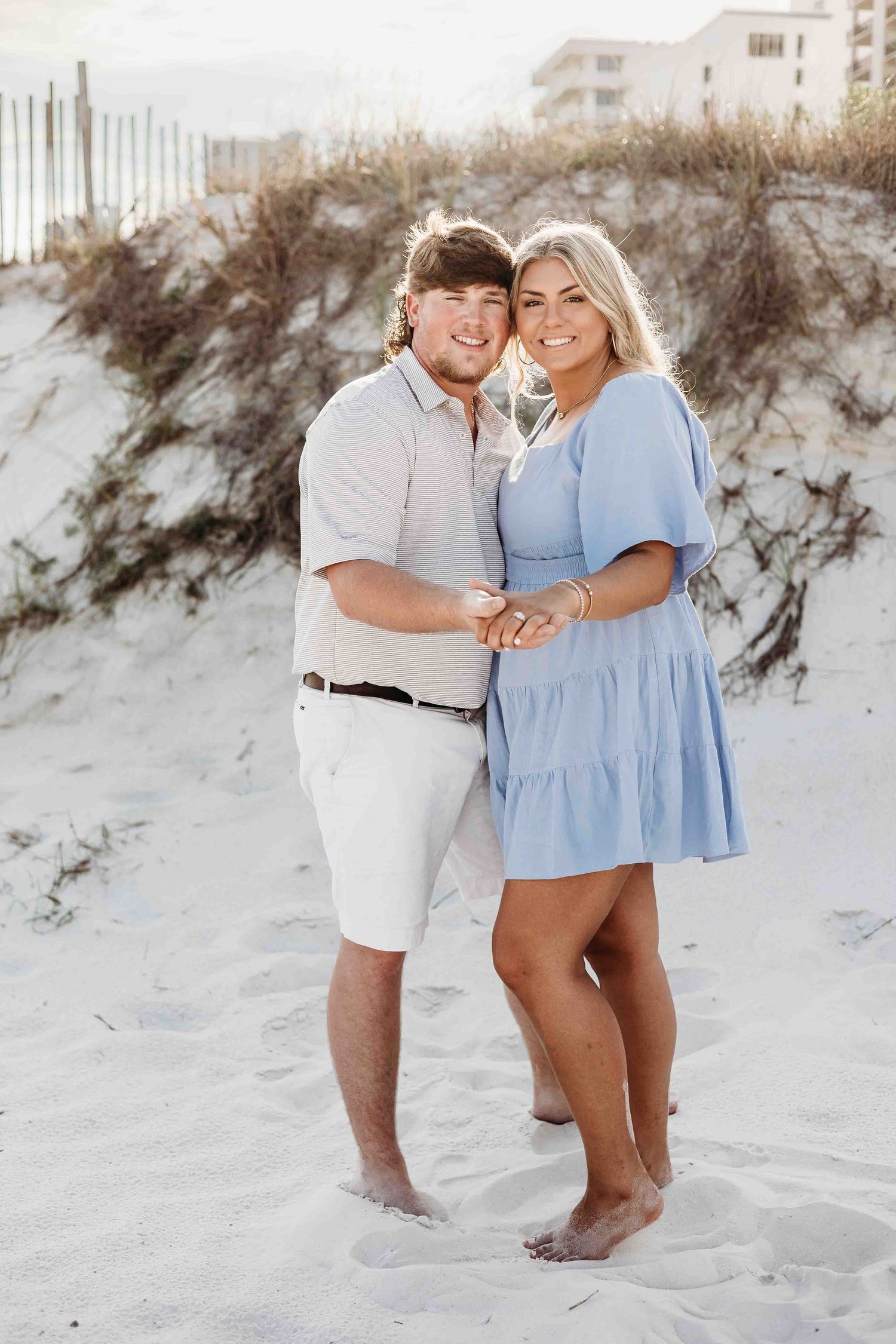 A romantic beach photoshoot featuring a couple in light blue and white outfits on sandy dunes.