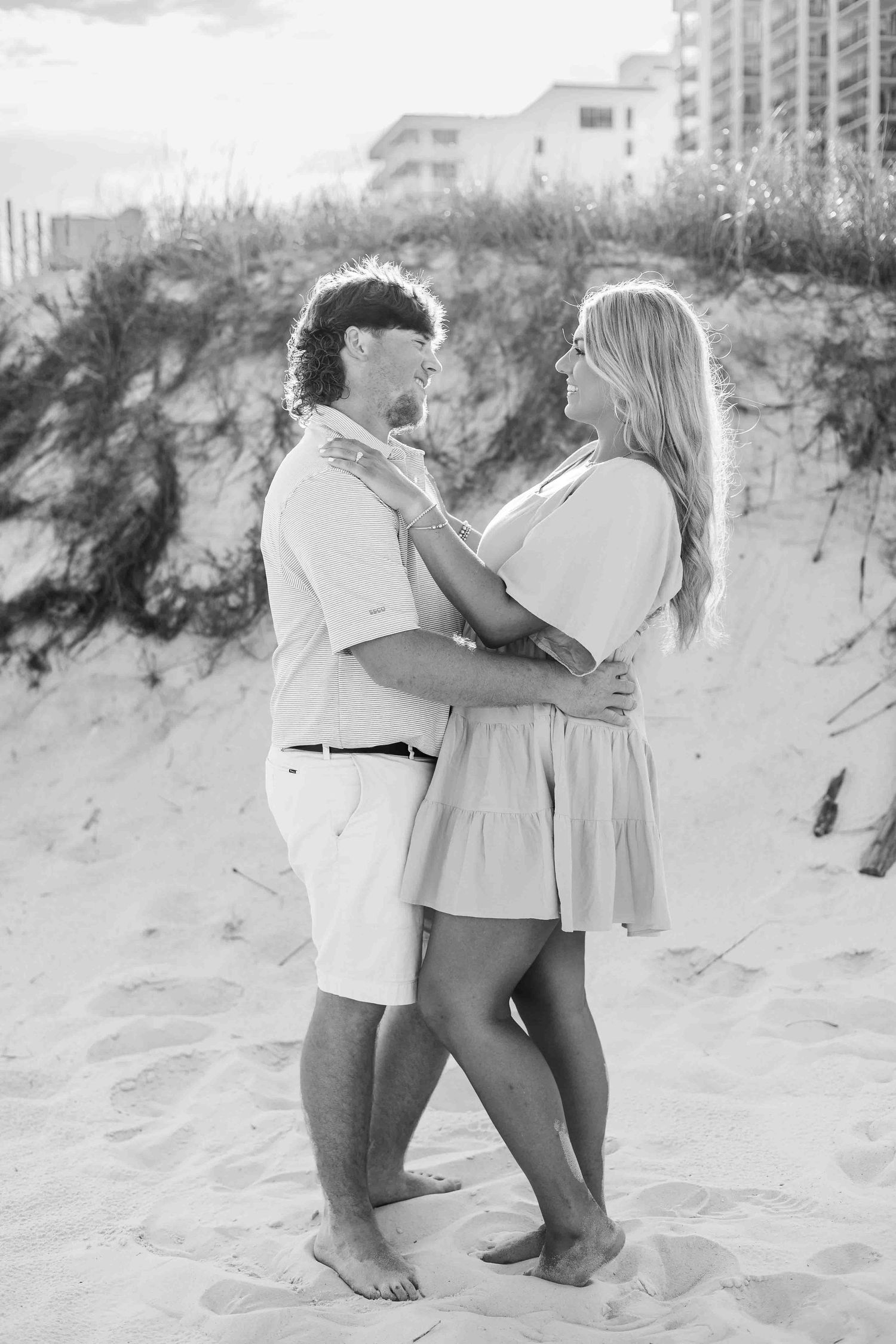 A black and white photo captures a tender moment between two people on sand dunes.
