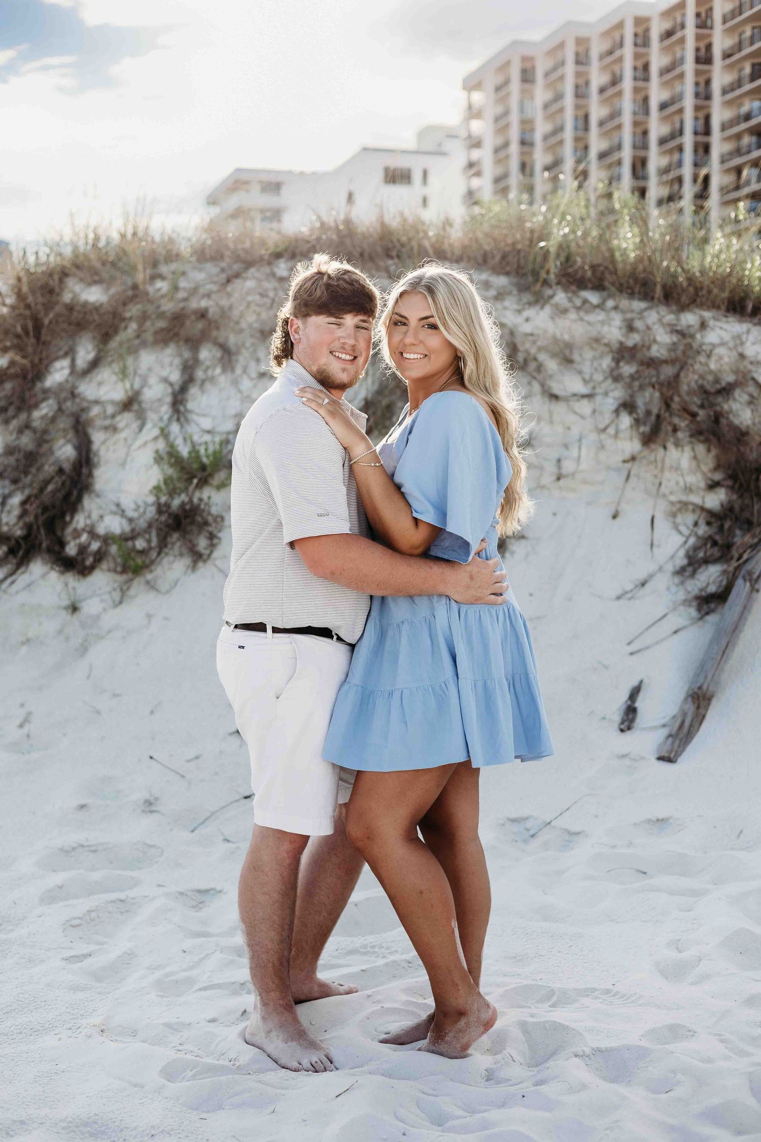 A couple poses together on white sand with coastal condominiums visible in the distance.