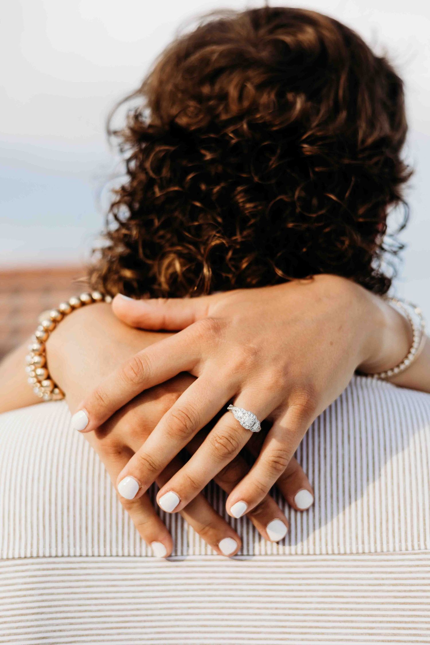 Close-up of hands wearing pearl bracelet and engagement ring resting on a light gray couch.