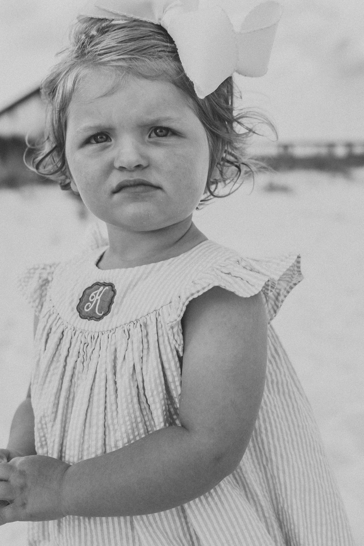 Close-up black and white portrait of a young child in a ruffled dress on the beach.