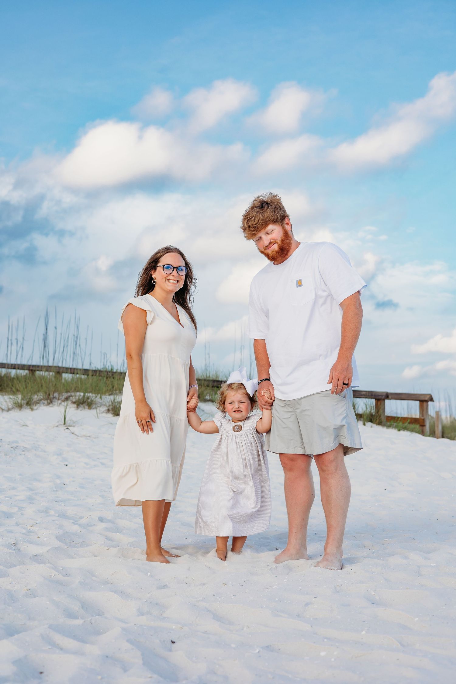 Three people in coordinated white and beige outfits stand together on a sunny beach.