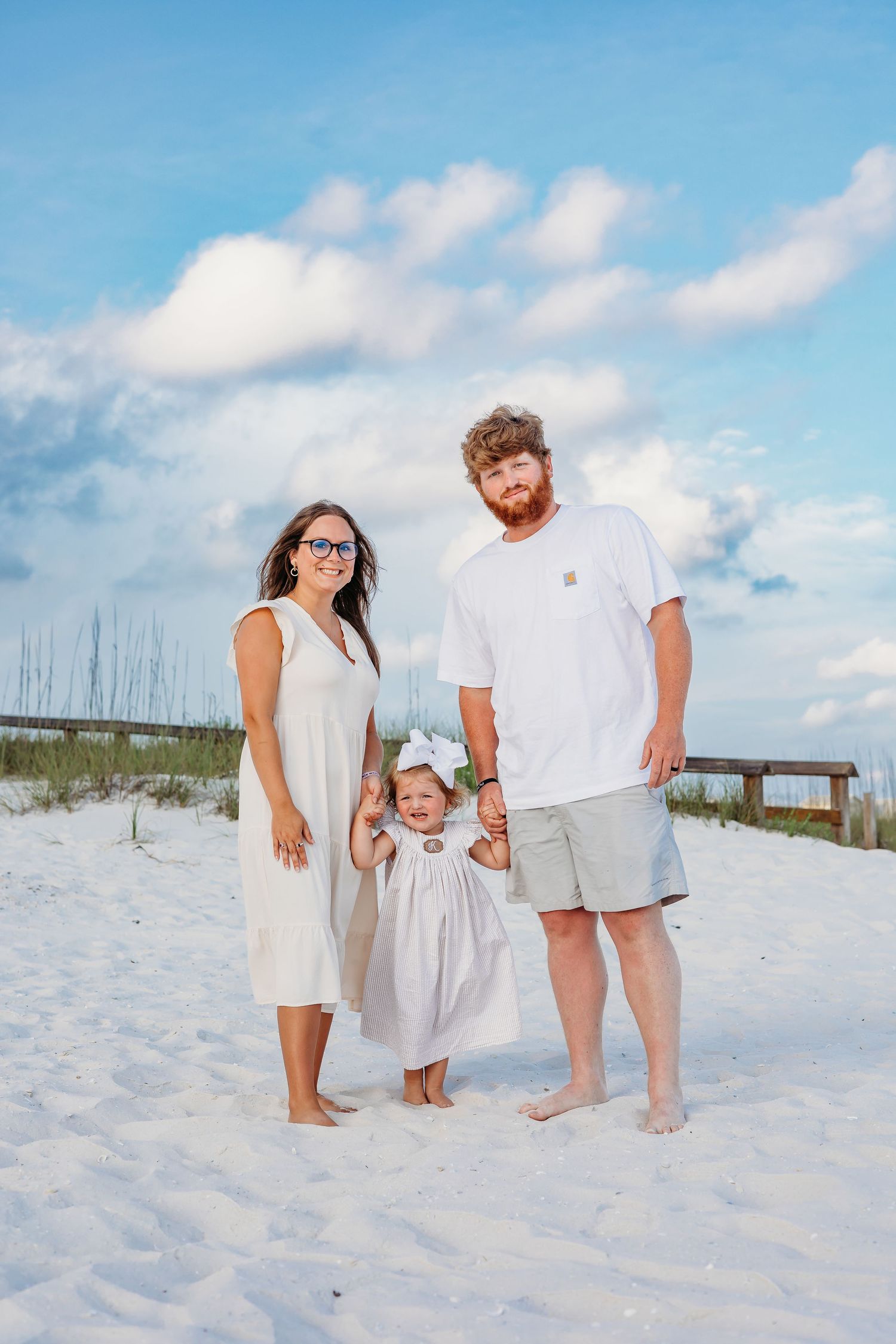 A family group takes casual photos while standing barefoot in white sand near beach grass.