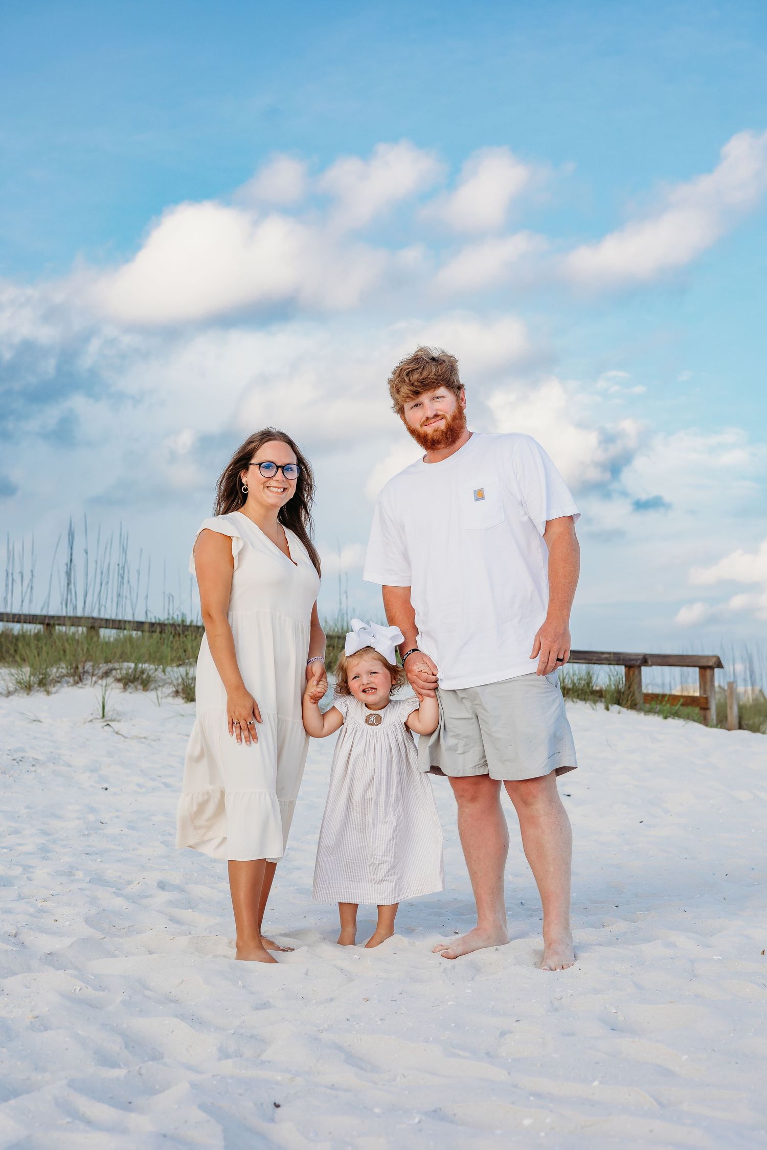 Three people in light colored clothing share a candid moment on a bright sandy beach.