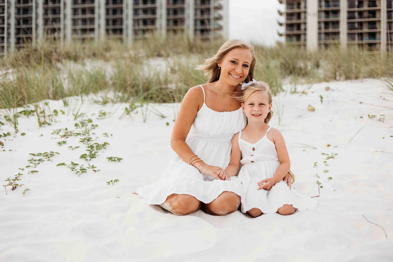 Mother and daughter in white dresses sitting together on sandy beach near condominiums.