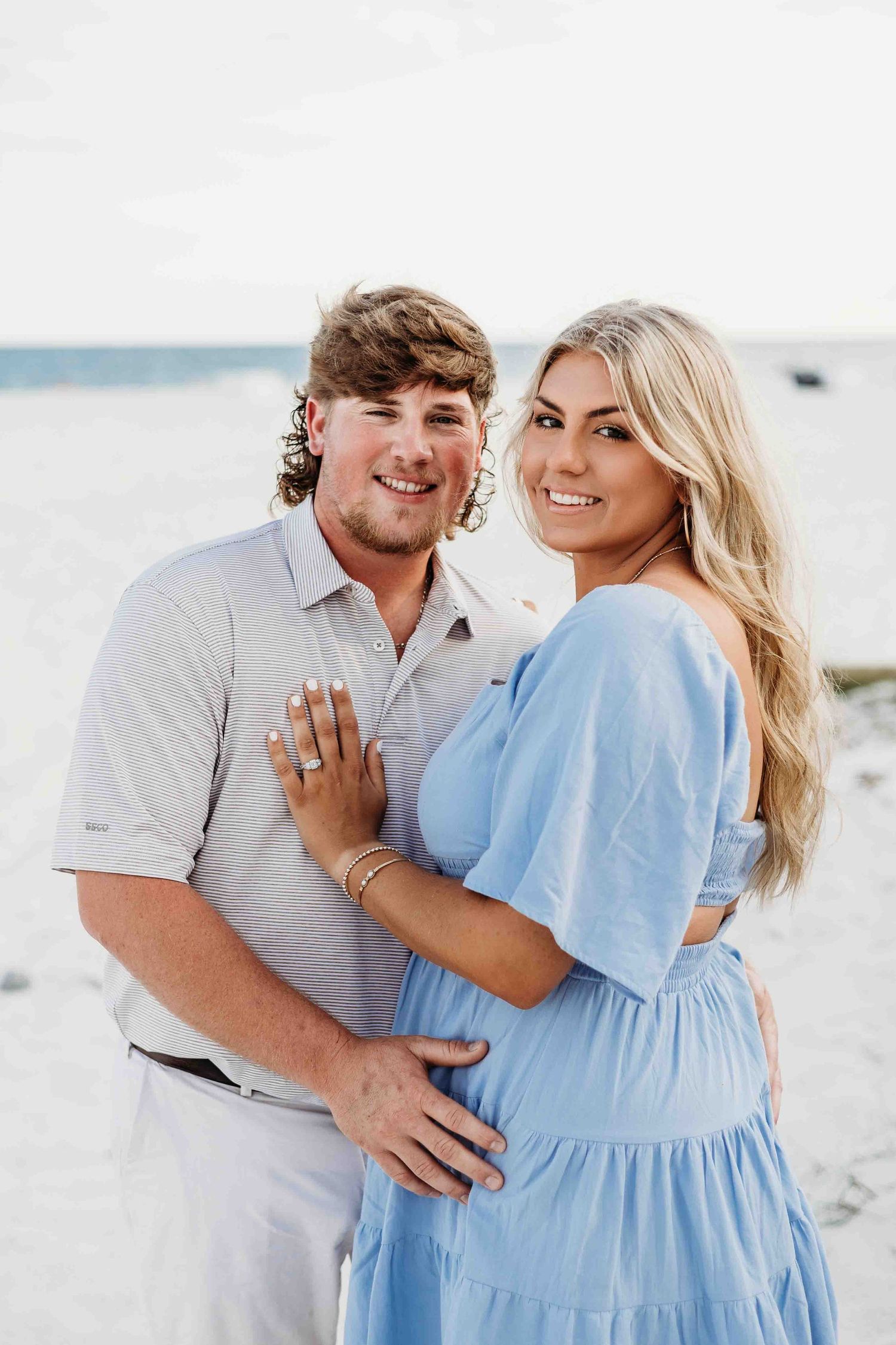 Newly engaged couple in casual attire showing off ring on sunny beach.