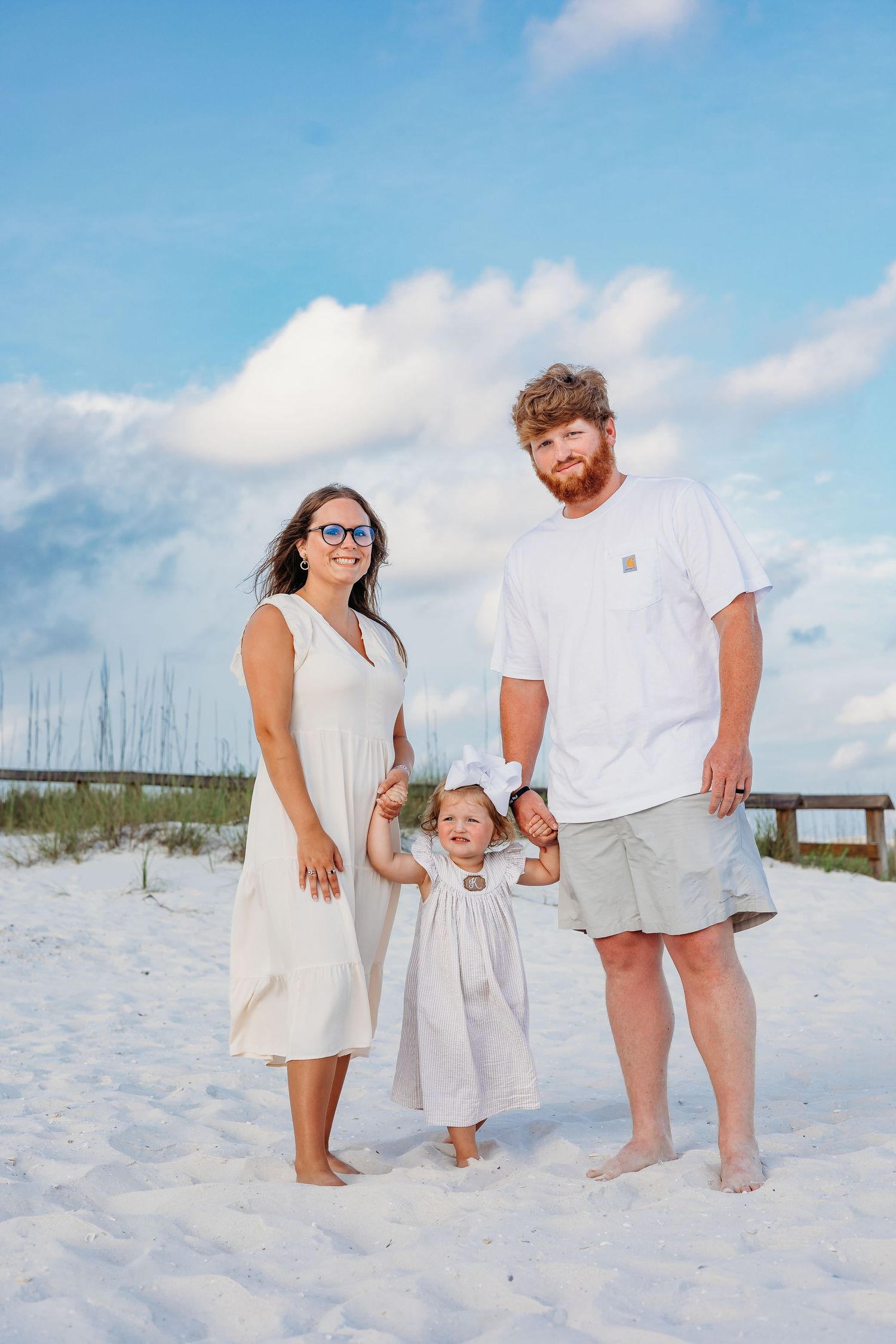 Family members in matching white outfits pose together on a sunny beach with dunes in the background.