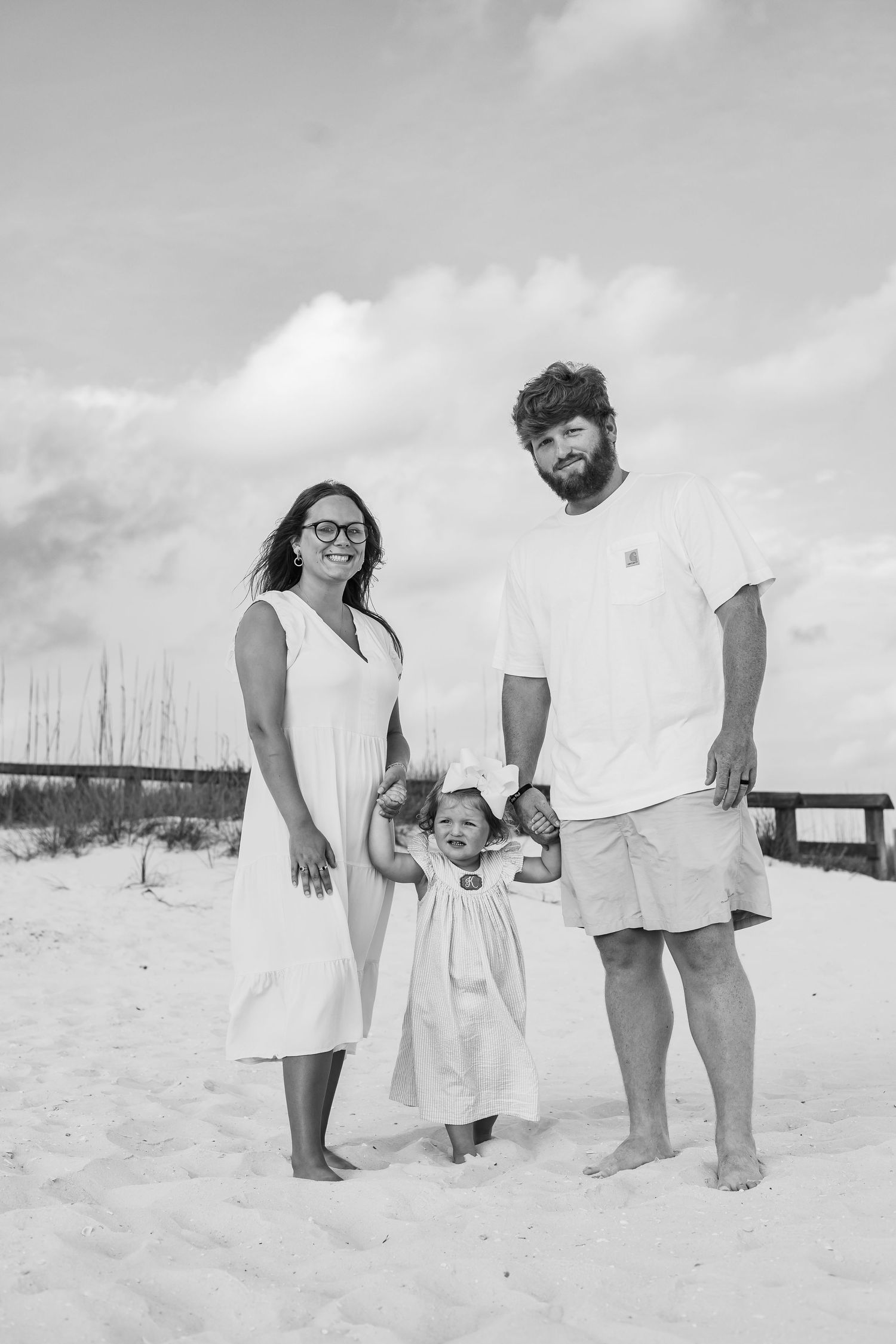 A black and white beach portrait of a family group standing in soft sand near dune fencing.