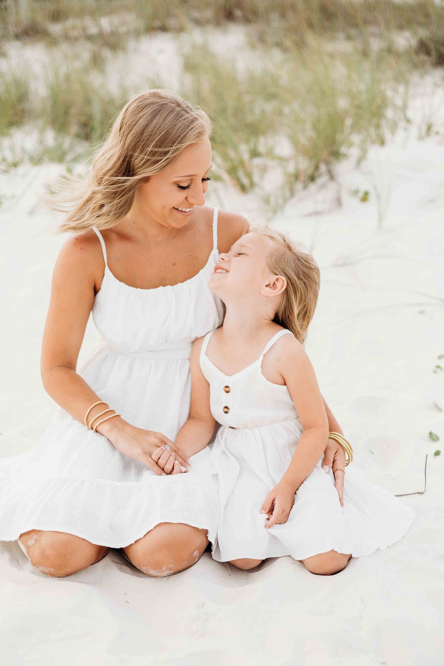 Mother and daughter in matching white outfits share a tender moment on sandy beach.