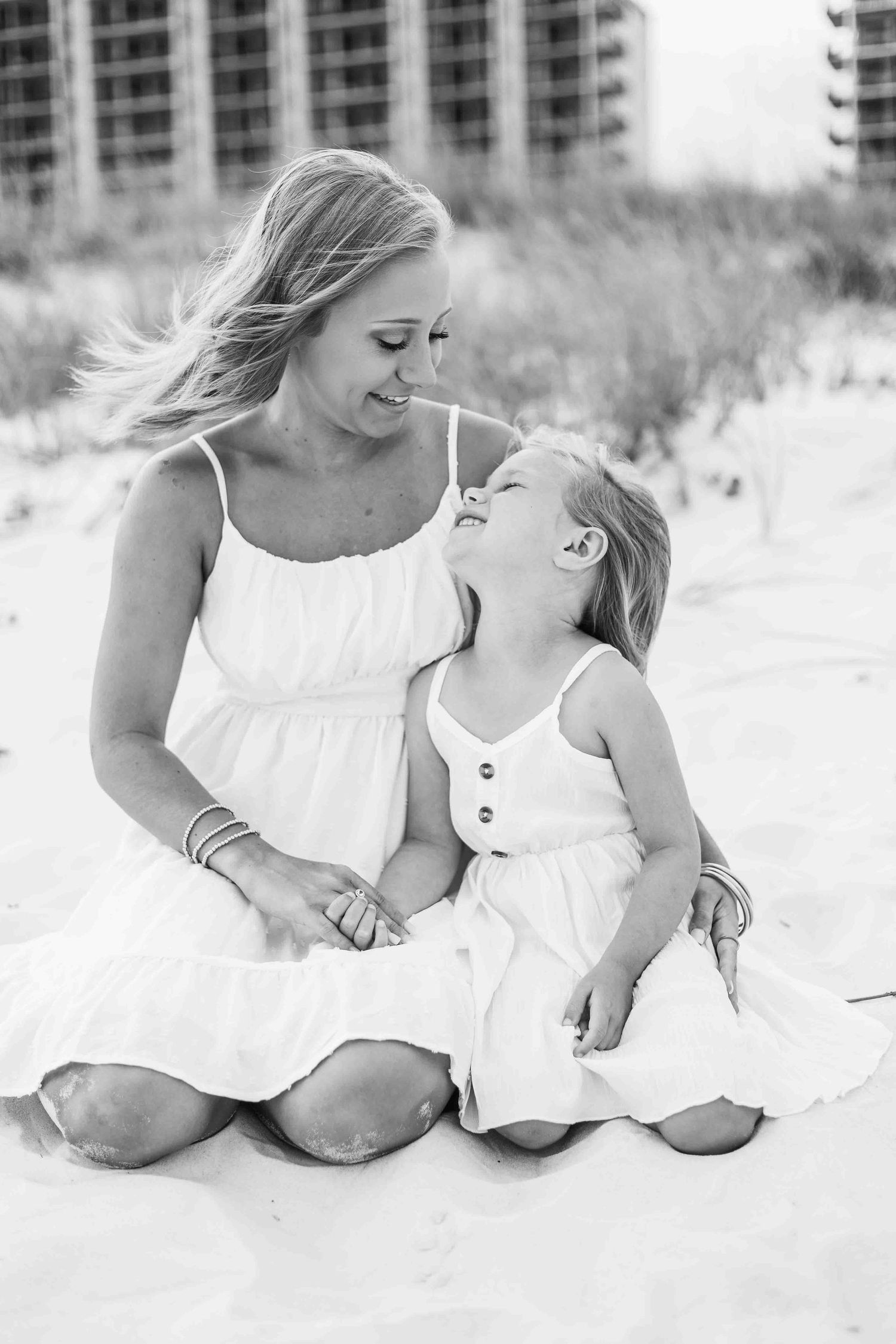 Mother and daughter in white dresses share tender moments together on a sandy beach.