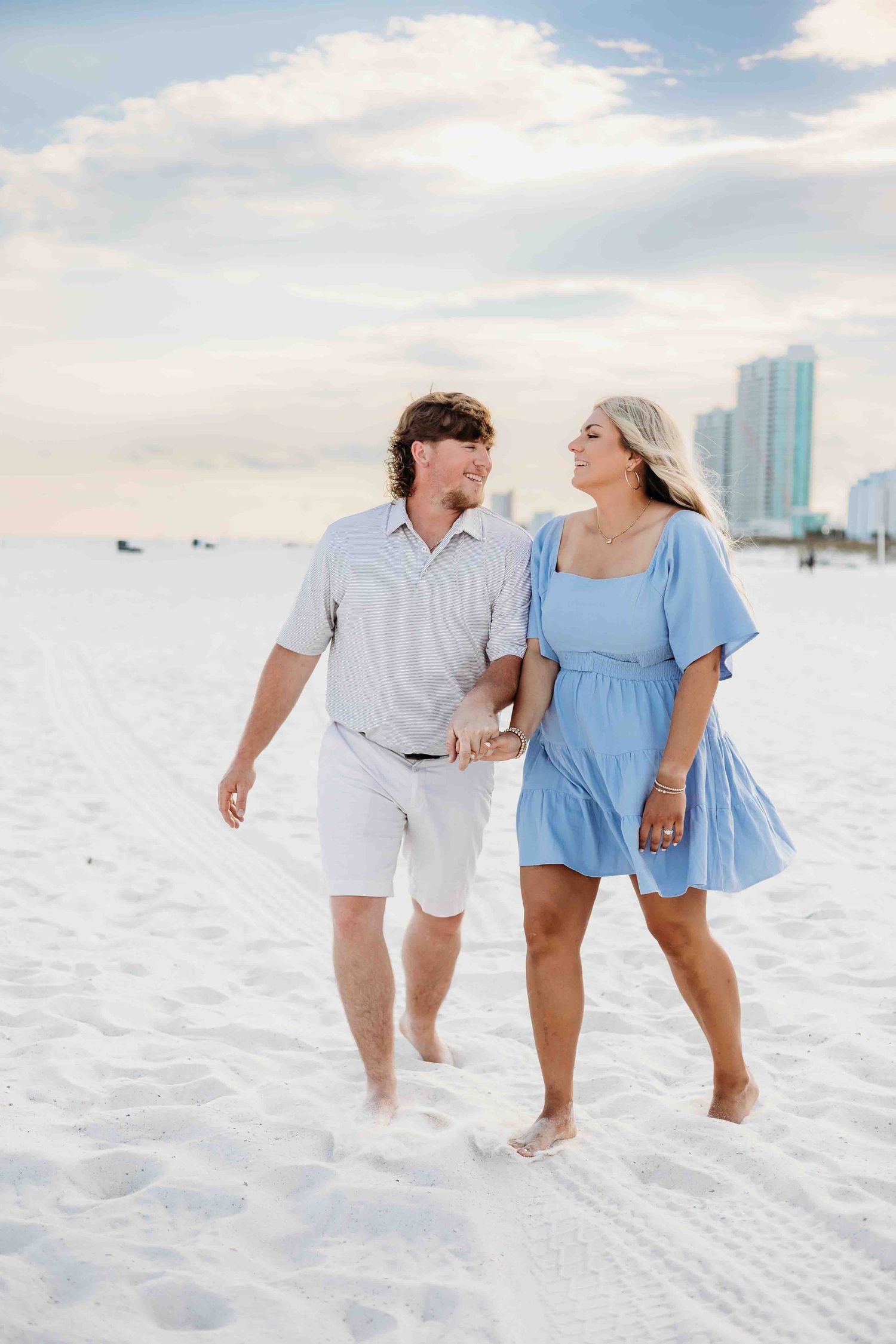Couple walking together barefoot on white sandy beach with coastal buildings in background.
