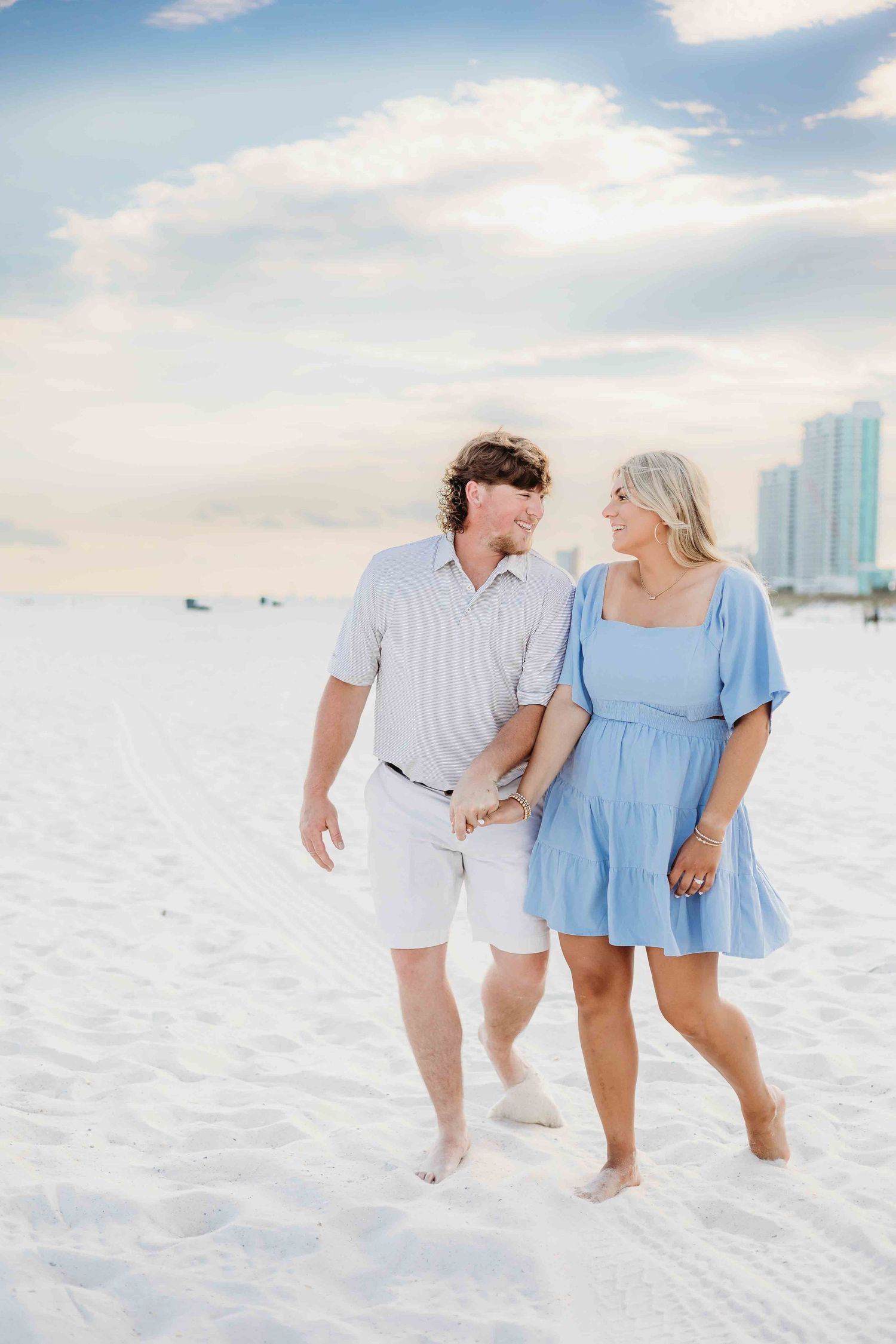 Candid shot of couple strolling along pristine beach with oceanfront buildings visible.