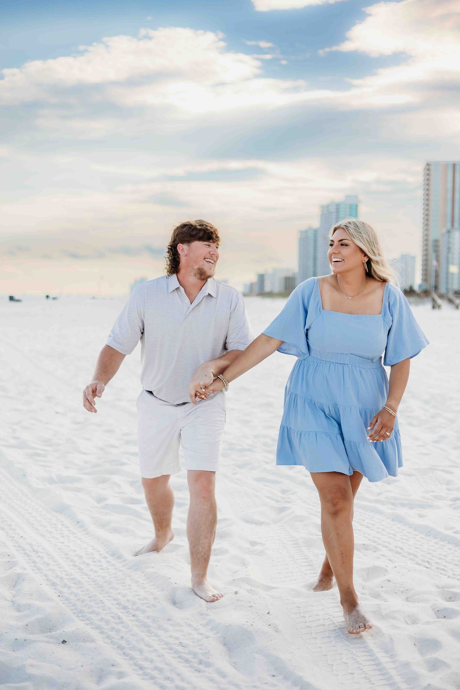 A couple strolls together on a pristine white sandy beach with coastal high-rises in the background.