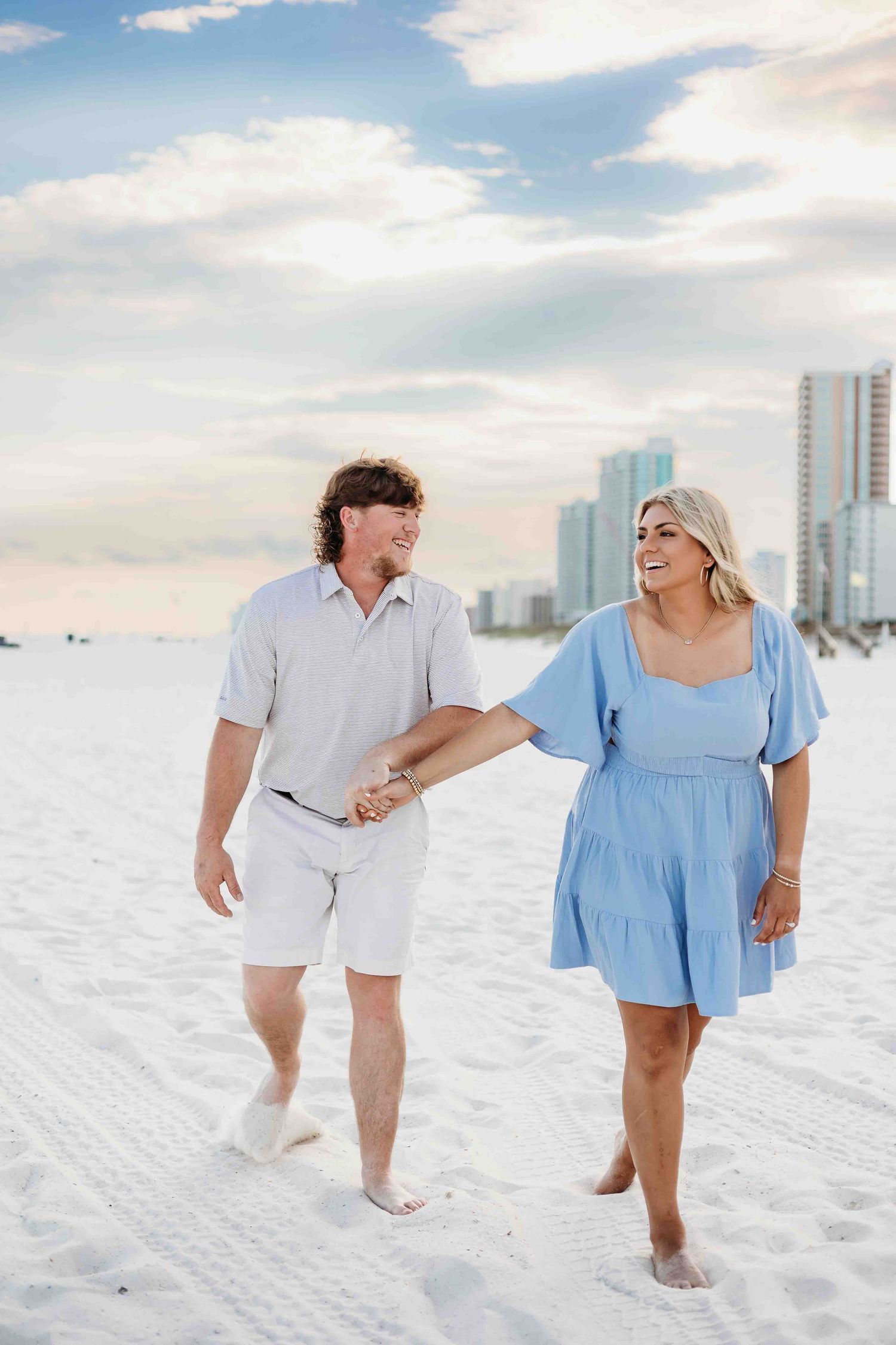 Two people walk barefoot along white sand beach near beachfront buildings on a sunny day.