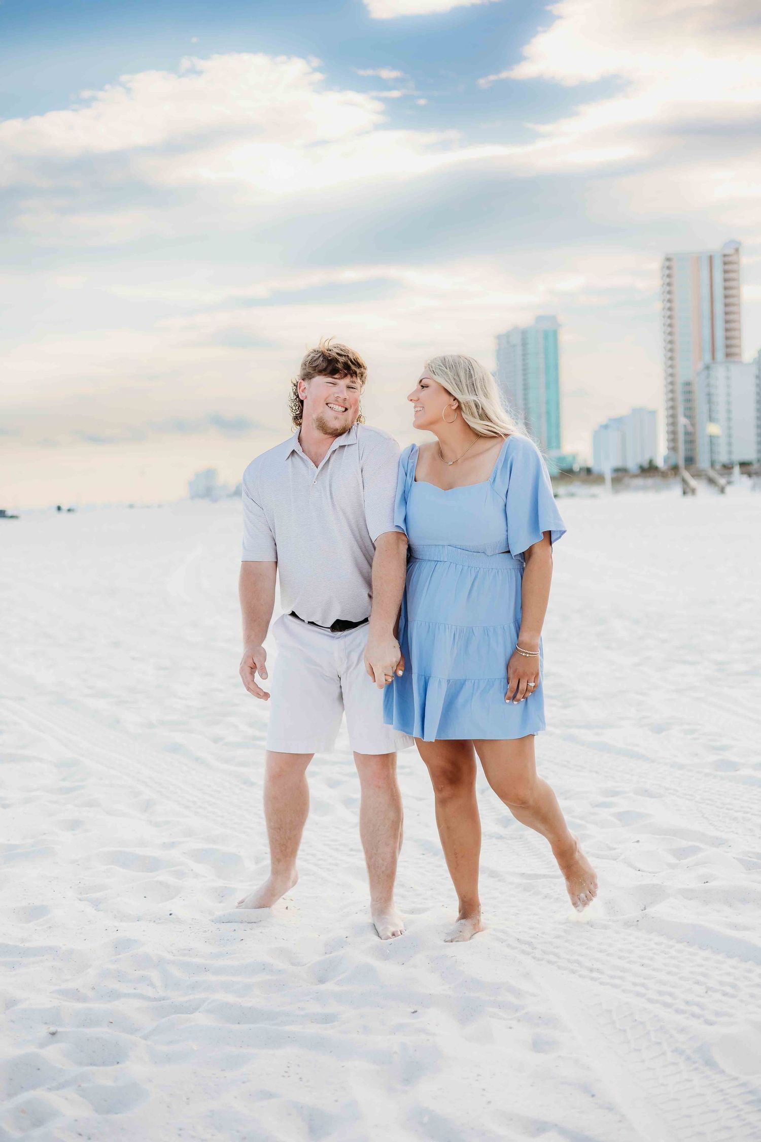 A romantic beach photoshoot featuring a couple walking through soft white sand near oceanfront towers.