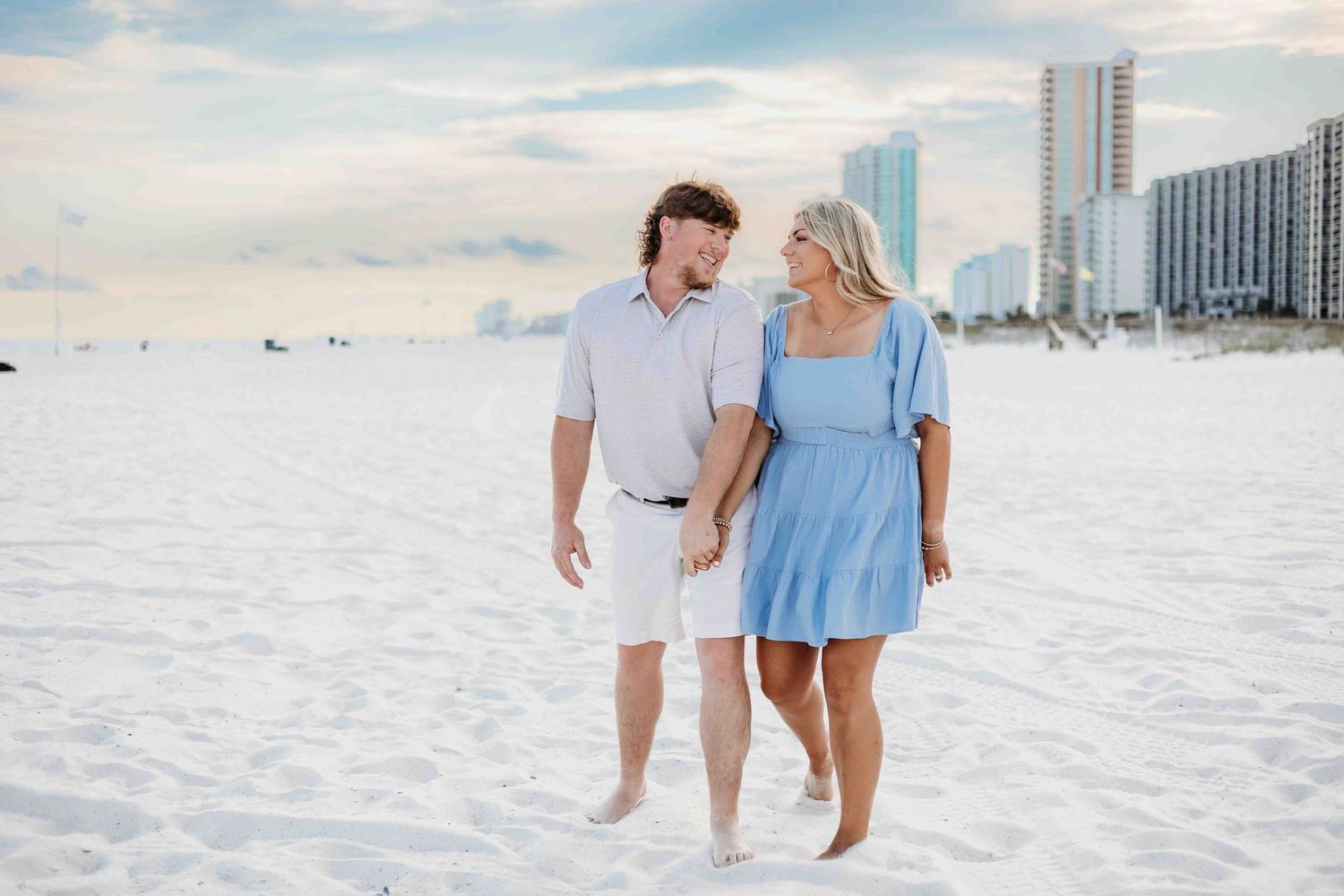 A couple enjoys a casual walk along a serene white sand beach with city skyline in the distance.