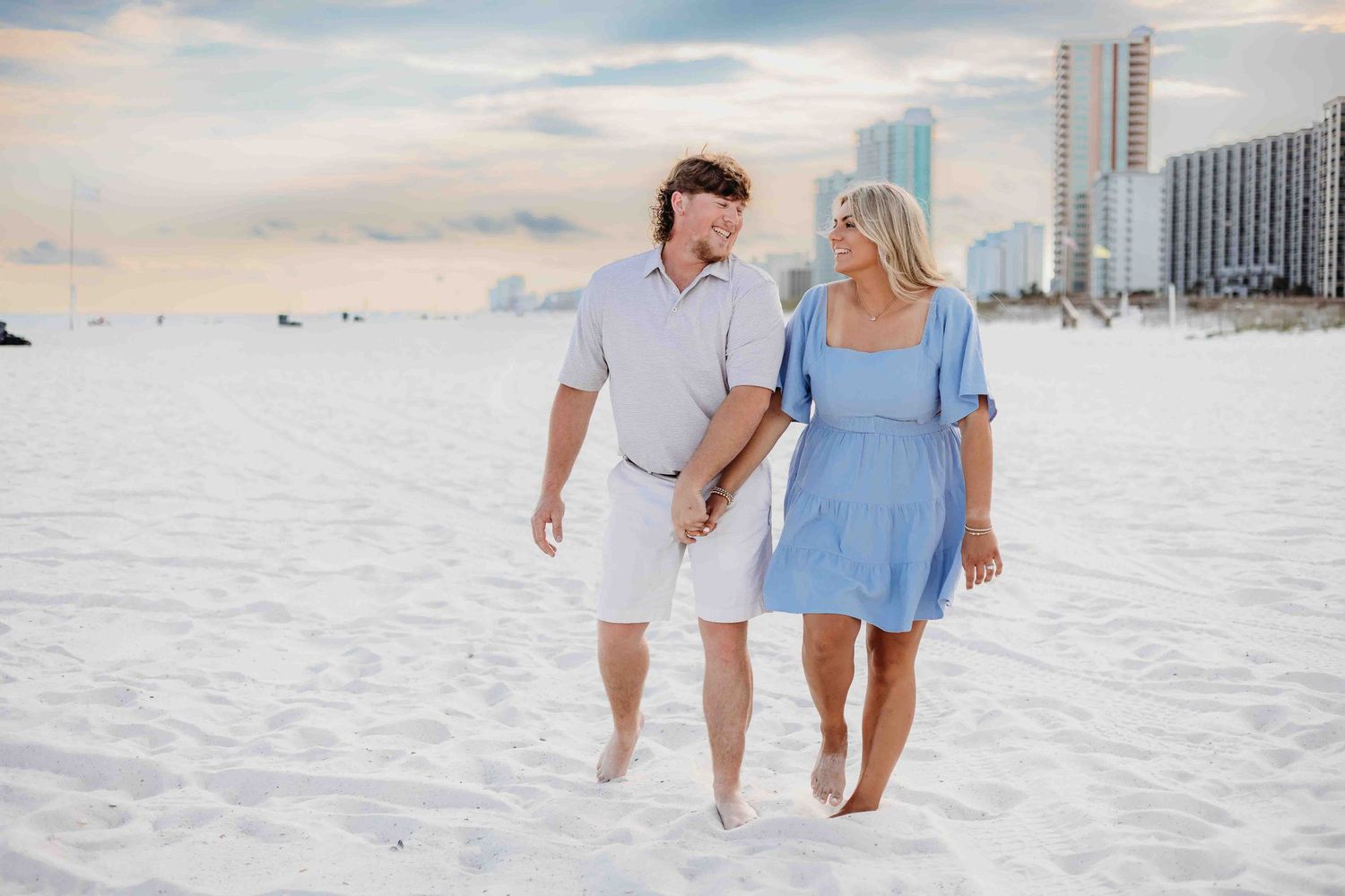 Two people share a romantic moment while walking on an empty white sand beach near coastal buildings.