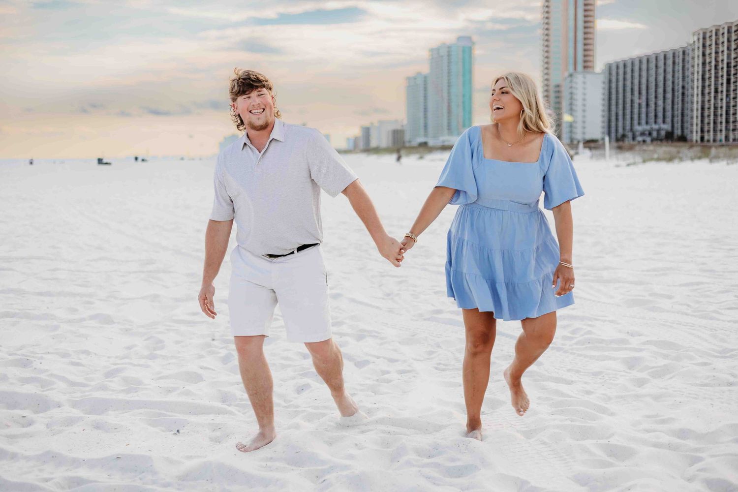 A couple holds hands while walking through pristine beach sand with high-rise buildings in view.
