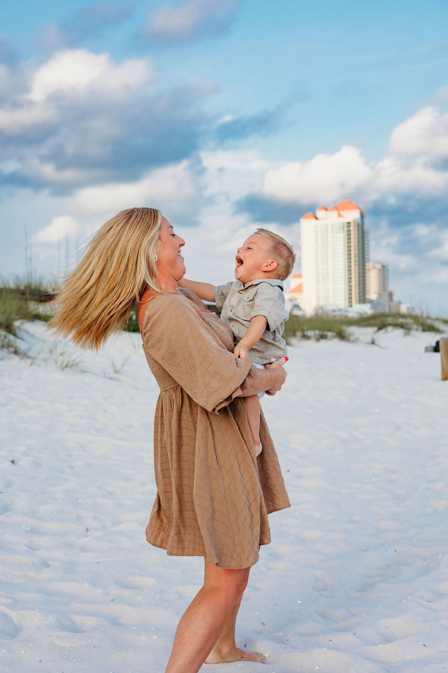 A tender moment between parent and child laughing together on a sunny beach with a high-rise in the background.