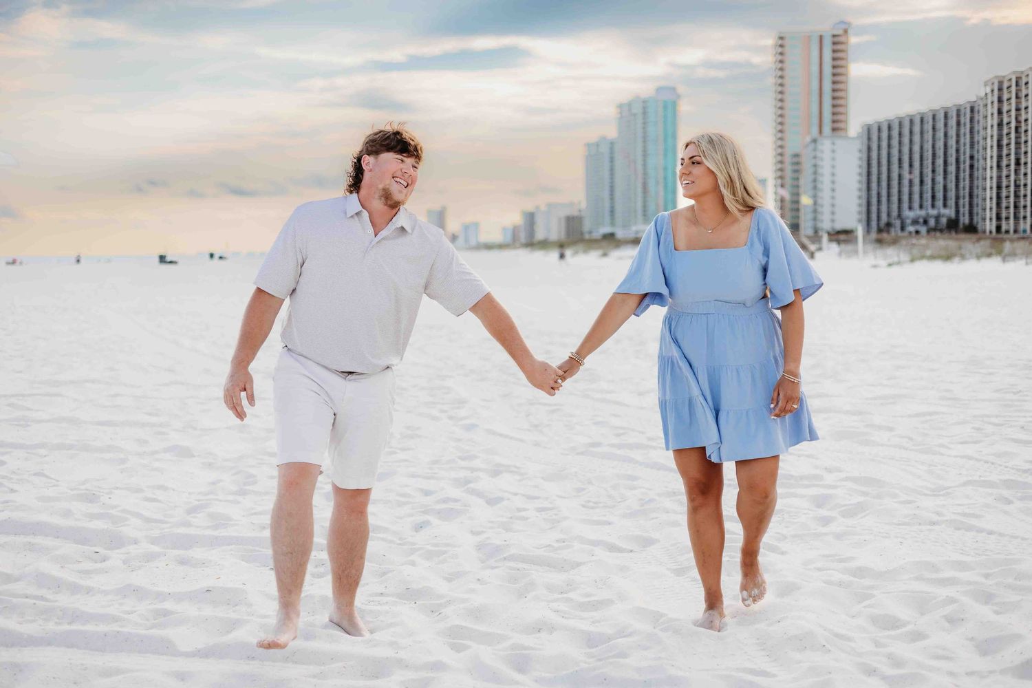 Two people walk hand in hand across a white sandy beach with coastal architecture in background.