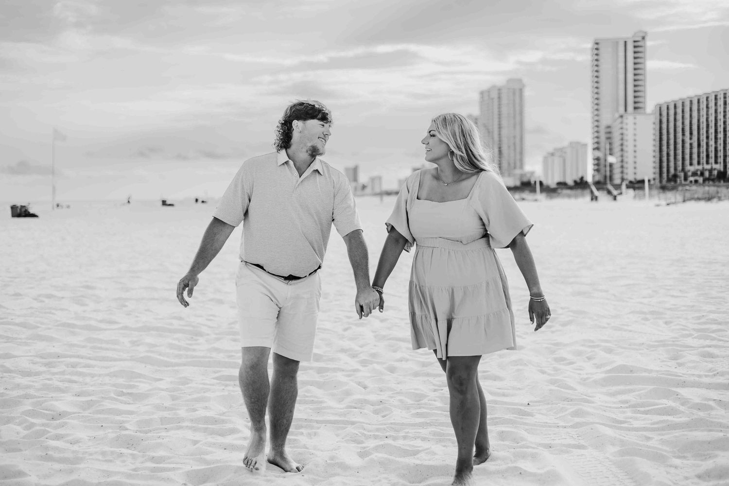 A black and white photo of a couple walking together on a beach with buildings in the distance.