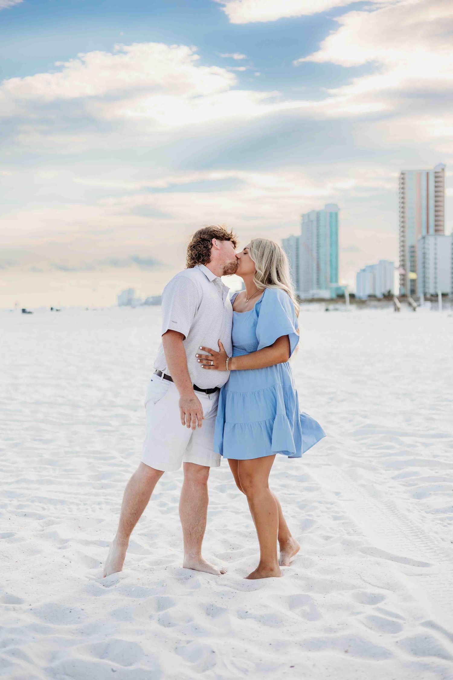 A couple embraces while walking along a sunny white sand beach with coastal skyline visible.