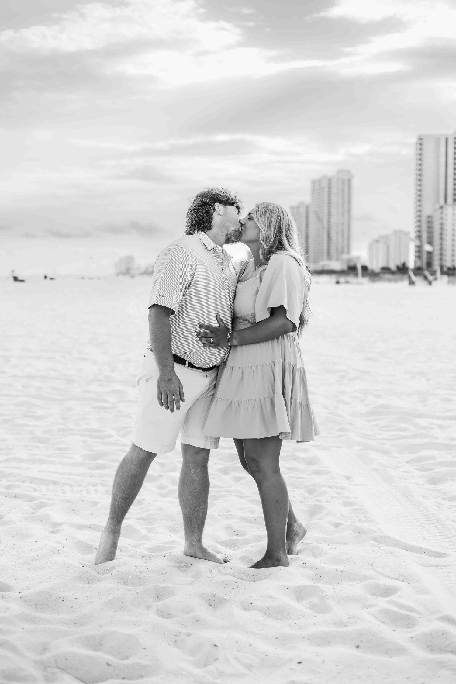 A black and white image captures an intimate moment between two people on a sandy beach.